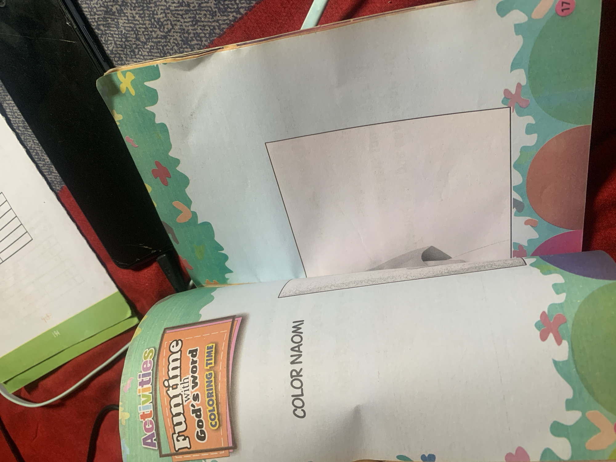 A close-up, overhead shot shows an open coloring book on a red textured surface, possibly a blanket or fabric. The book is open to a page with the title "COLOR NAOMI" in large, simple text. To the left, a colorful header reads "Activities Funtime with God's word COLORING TIME." The right page of the book features a large, empty rectangular box, likely a space for a drawing or coloring activity, bordered by a whimsical design of green, blue, and pink shapes. The bottom edge of both pages is decorated with a border of colorful, abstract shapes resembling flowers and rolling hills. A small red circle with the number "17" is visible on the bottom right corner of the right page. The background shows a portion of a green folder and a black electronic device. The image does not contain any information that can be used to determine its location in Jauro Yinu, Nigeria, nor does it depict any people, actions, or interactions.
