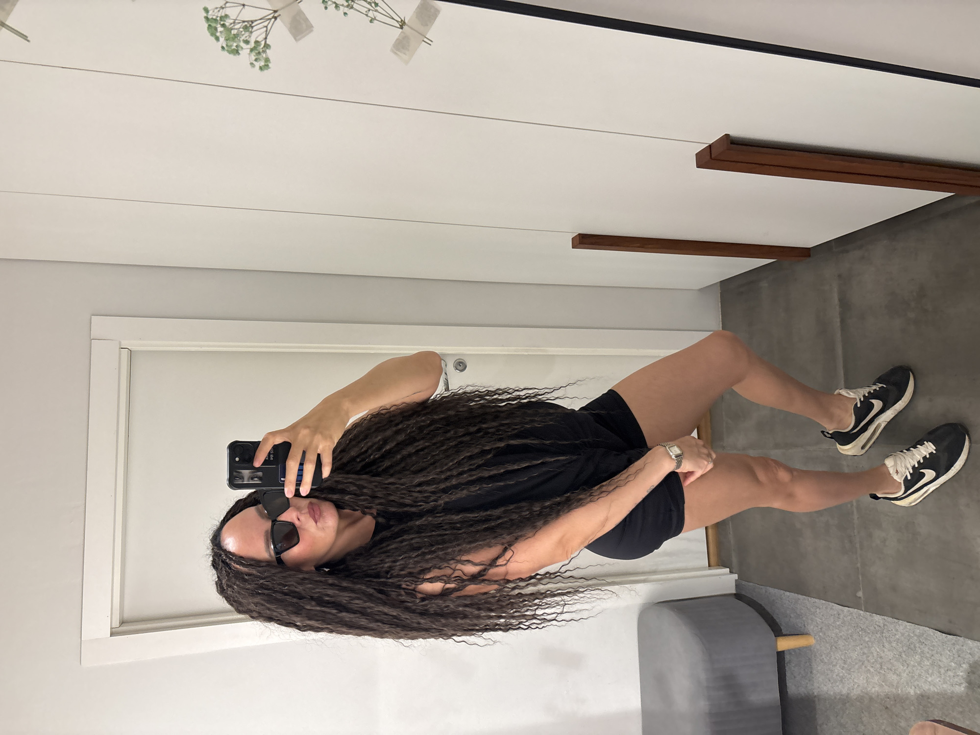 A woman with long, dark, curly hair is taking a selfie in a room. She is wearing sunglasses, a black t-shirt, and black shorts, and is holding a black smartphone in her right hand. She is standing and facing the mirror, which reflects a white wall with wooden decorative elements. The floor appears to be concrete, and there's a gray ottoman-like object in the lower-right corner. Her legs and shoes are visible; she is wearing black and white sneakers. The location cannot be ascertained from the image and is not necessarily Chaikovsky, Russia.