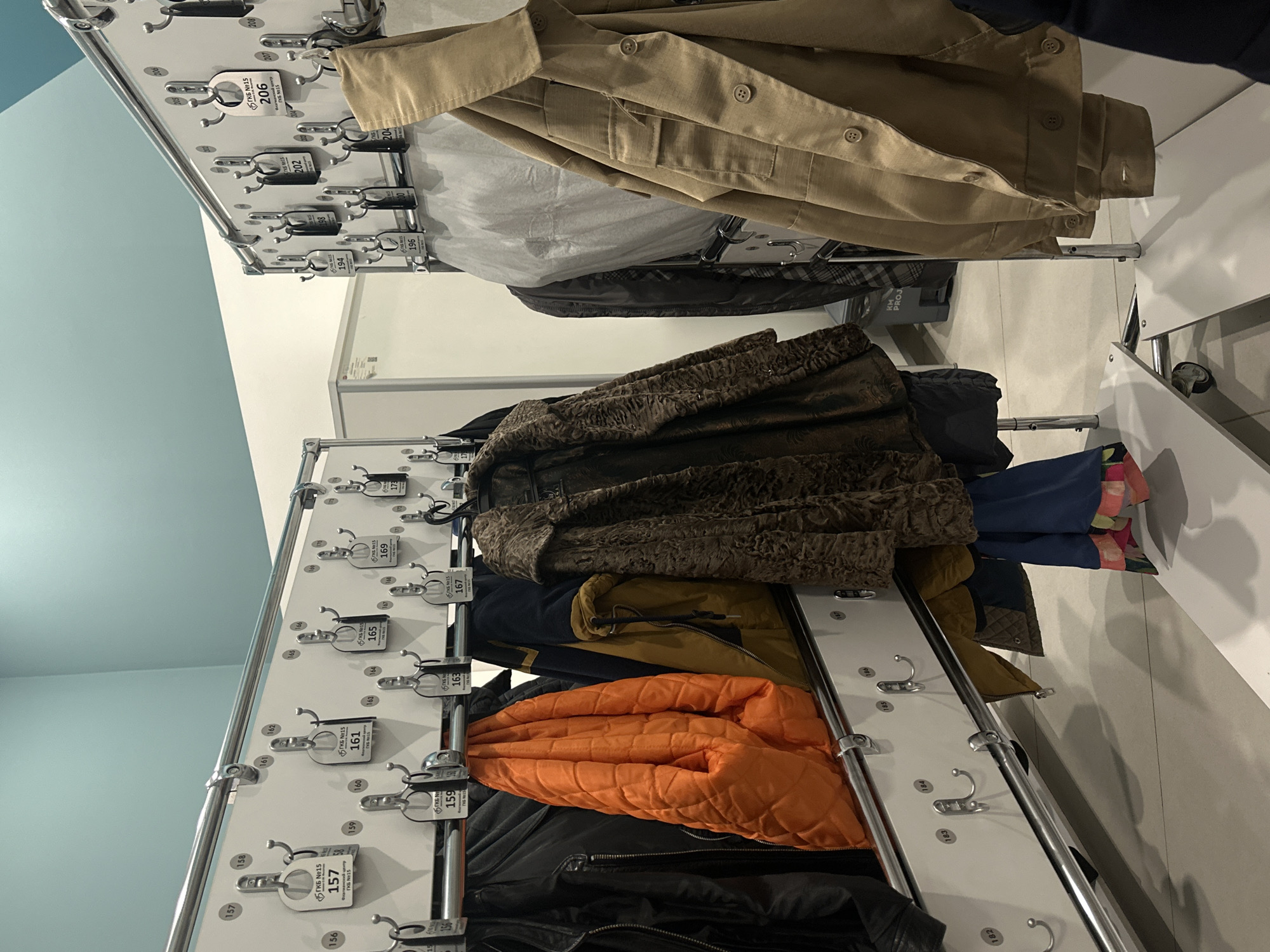 This indoor shot shows two double-sided coat racks, likely located at Flagship Center TKS No. 15 in Moscow, Russia, as evidenced by the tags.
The top coat rack features a tan button-down shirt, a light gray plastic bag, and a dark jacket.
The lower rack displays coats in various colors and textures, including a brown fur coat, navy jacket, mustard jacket, and orange quilted jacket. A black leather jacket hangs at the bottom. Each coat is hung on a hook with a numbered tag.