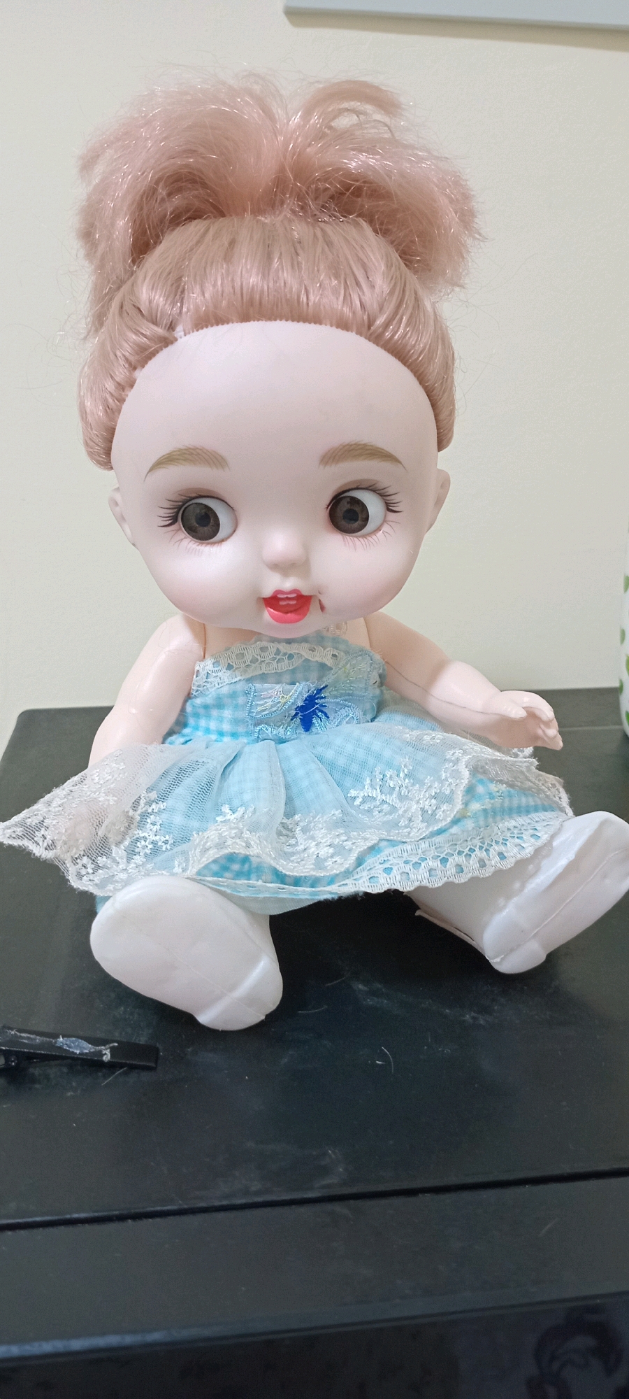 The image shows a close-up, frontal view of a doll with light pink hair styled in two messy buns. The doll has large, wide-open brown eyes with long eyelashes, small eyebrows, and red lips. It is wearing a light blue and white checkered dress with white lace trim and a blue decorative element in the center of the bodice. The doll's arms are slightly out to the sides, and its white feet are visible. It is sitting on a dark, flat surface that appears to be a table or shelf. The background is a plain, light-colored wall. There is a black object, possibly a clip or fastener, on the dark surface in the lower left corner of the image. The scene does not provide any specific visual cues to indicate its location within the city of Altynkul, Uzbekistan.