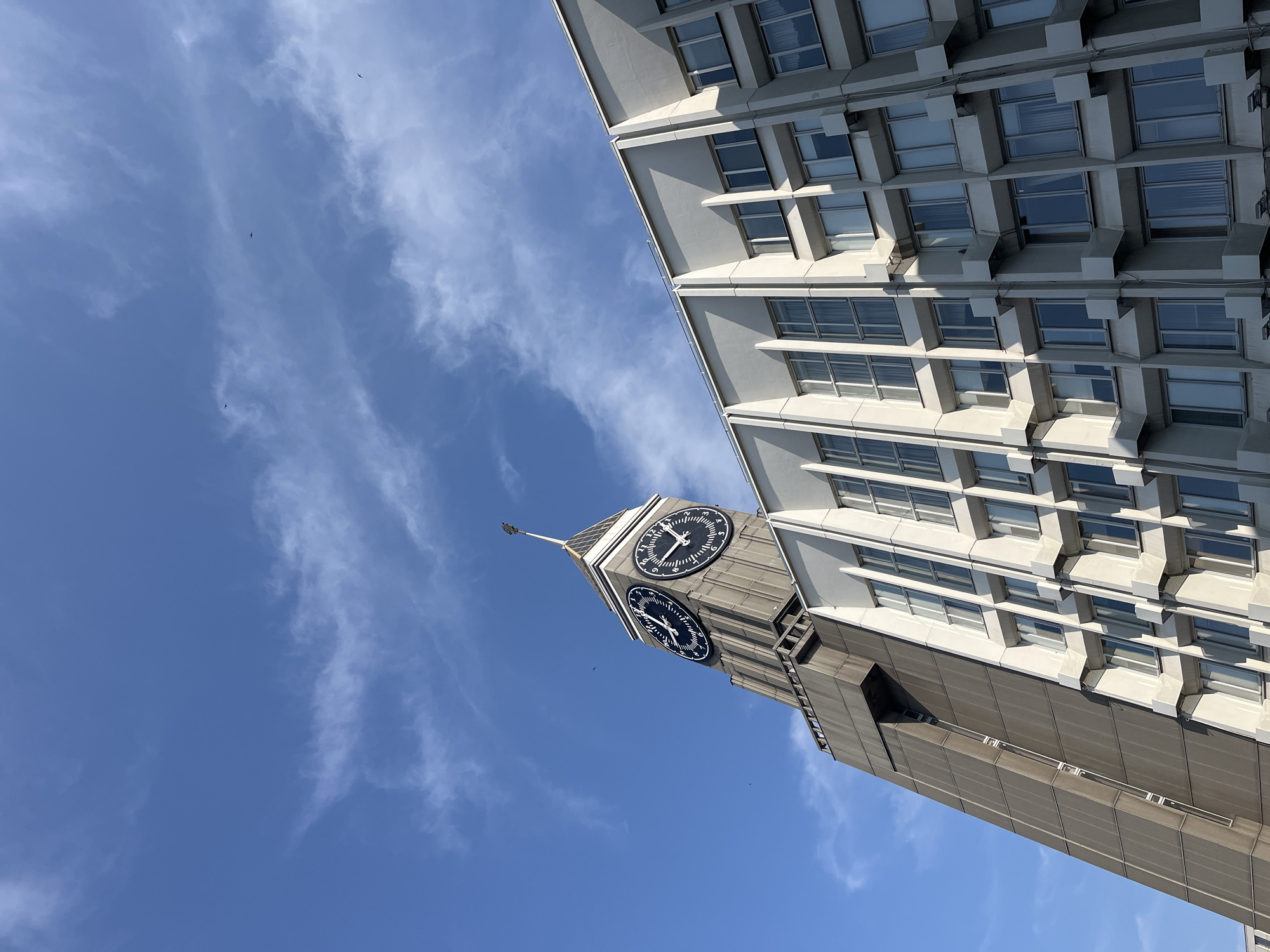 The image captures a low-angle view of a distinctive building in Krasnoyarsk, Russia, featuring a prominent clock tower. The sky is a clear blue, with scattered wisps of white clouds. Several small birds are visible in the sky, appearing as dark specks against the blue.
The building itself has a striking architectural design. The left side of the image is dominated by a tall, grey, angular tower that houses two large circular clocks with black faces and white numerals. The clock hands indicate approximately 10:40. The tower is topped with a pointed spire and what appears to be a weather vane.
Adjacent to the clock tower, and forming the main facade of the building, is a lighter-colored structure characterized by a repeating pattern of windows. These windows are recessed and framed by geometric architectural elements, creating a textured, modern appearance. The sunlight casts shadows on the facade, highlighting the depth and angles of the design. The building has multiple stories, and the windows are arranged in vertical and horizontal rows. Some of the windows show reflections of the sky. No people are visible in the image. The overall impression is one of urban architecture set against a bright, open sky.