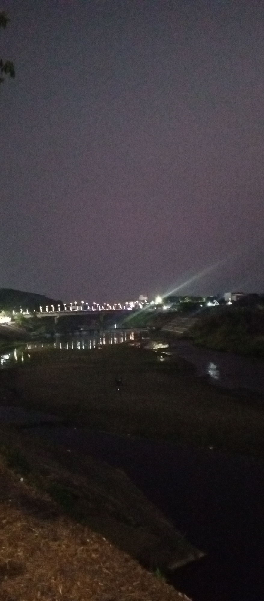 The image captures a nighttime scene of the Madiun River in Madiun, Indonesia. A long bridge, illuminated by numerous lights, spans across the river in the background, with reflections of these lights shimmering on the water's surface. Buildings and what appear to be streetlights are visible on the riverbanks and beyond the bridge, suggesting an urban or semi-urban environment. A bright beam of light, possibly from a spotlight or a vehicle, cuts across the sky on the right side of the image. The foreground shows the dark, still water of the river and the earthen bank on the right, with some dry vegetation. The overall atmosphere is dark and slightly hazy, typical of a night scene in a populated area.