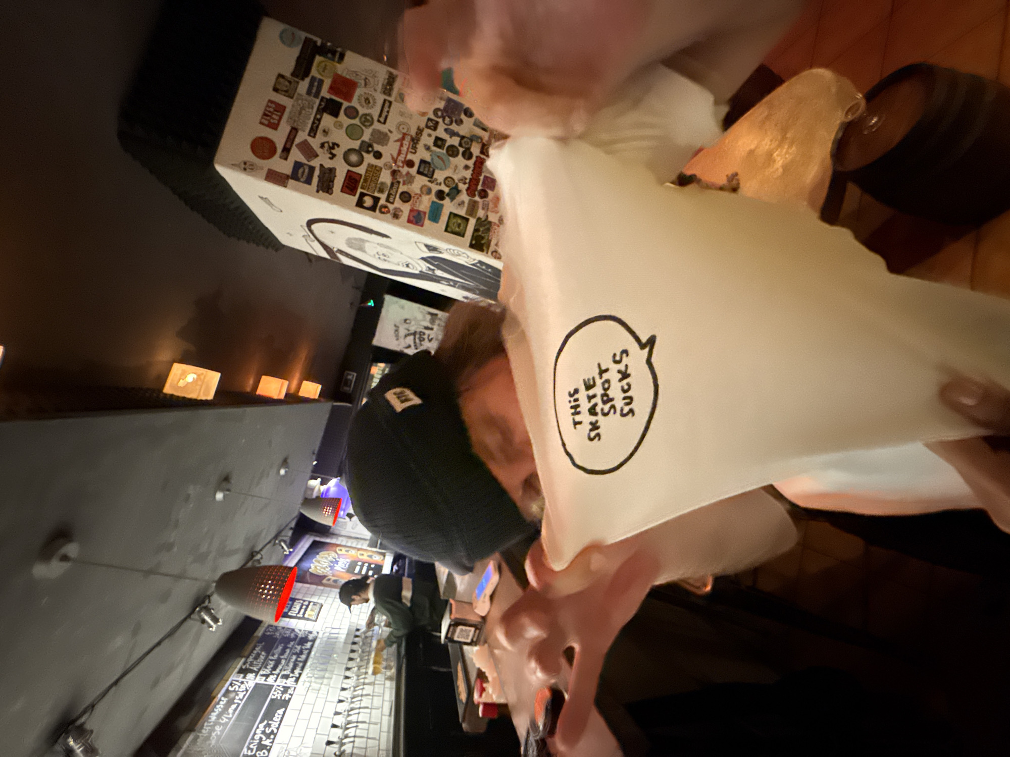 The image appears to be taken inside a bar or restaurant in Barcelona, Spain. A person is holding up a white bag with the text "THIS SKATE SPOT SUCKS" printed in a black speech bubble on it. Other people are standing behind a counter or bar. The bar menu is visible in the background. The location seems to be a modern, somewhat trendy establishment.