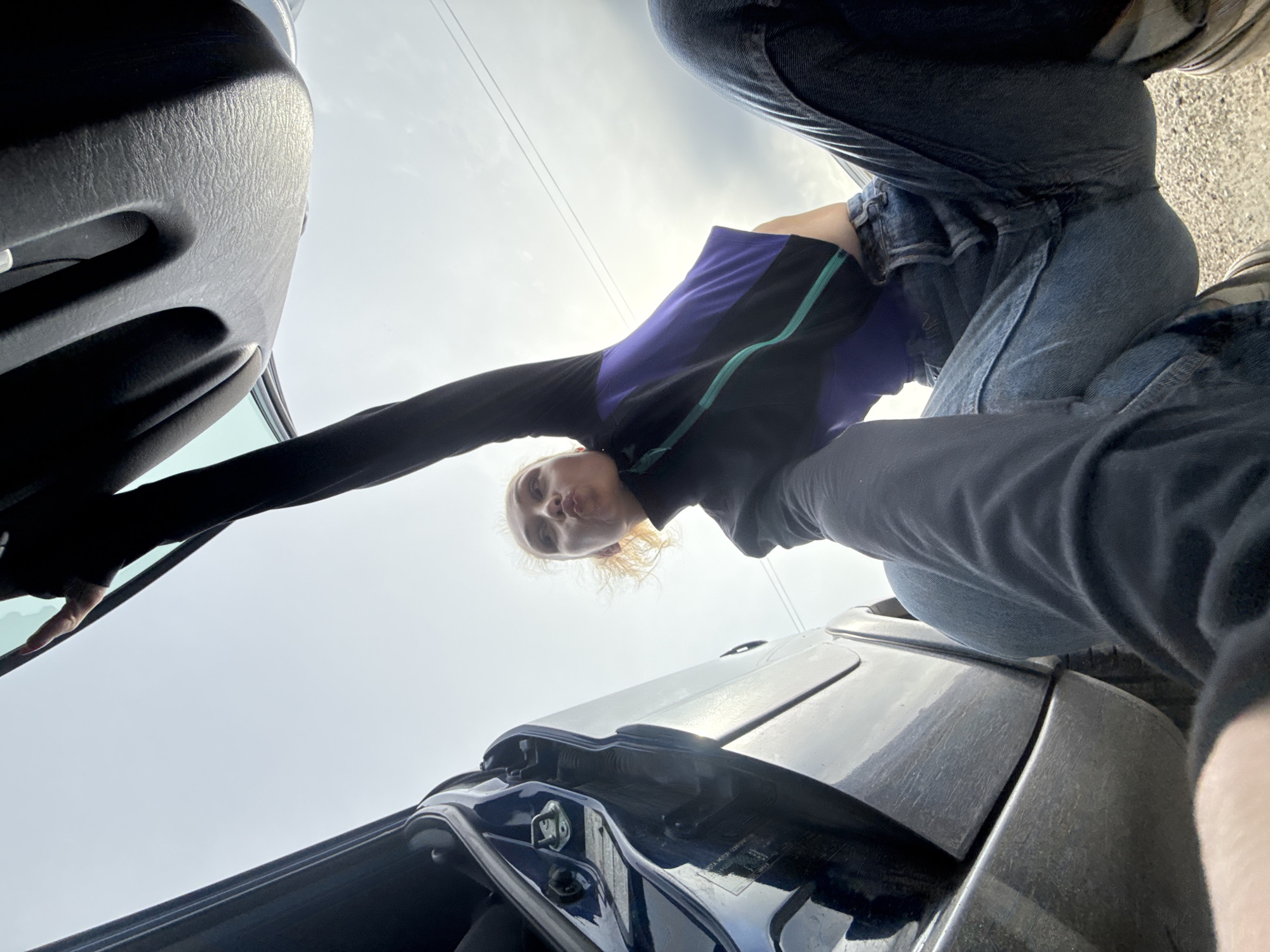 A person with fair skin and light blonde hair is seen from a low-angle perspective, looking down at the camera. They are wearing a dark long-sleeved shirt with a blue and black torso section and a green stripe. The person is also wearing blue jeans. Above and to the left of the person, there is part of a car door, open. Below the person is the hood of a car, with the car's surface reflecting the sky. The sky is visible in the background. Based on the image, the location in the city of Chaikovsky, Russia cannot be determined. There is no indication in the image to point to that location.
