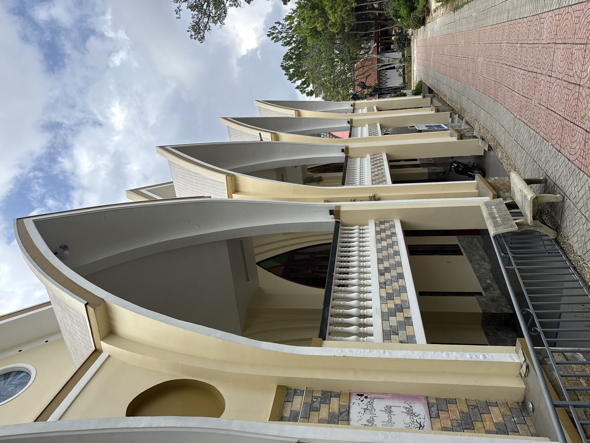 The image captures the distinctive side facade of a multi-story building, likely the Domaine de Marie Church in Da Lat, Vietnam, as indicated by a visible plaque. The building features light yellow walls contrasted with white and light grey trim. Its most striking architectural element is a series of repeating, upward-curving canopy-like structures or roofs, creating a wave effect along its length. Sections of the building are adorned with decorative square tiles in muted shades of grey, white, black, and beige, arranged in a mosaic pattern. White balustrades are visible along railings, and a round window with stained glass is seen on the left side of the structure.
To the right of the building, a neatly paved pathway extends into the distance, featuring a central section of reddish-pink rectangular tiles flanked by grey tiled sections. Lush green trees are visible in the background, mostly in the upper right. A dark-colored motorcycle is parked beneath one of the building's overhangs. A stone bench sits beside the pathway, and a metal railing or staircase descends near it.
The sky above is overcast with scattered white and grey clouds, suggesting a cloudy day. No people or specific activities are depicted; the scene is static.
Visible text includes:
- On the stone bench: "GÓC NGỒI CỦA CÁC ÔNG BÀ" (Vietnamese for "Sitting corner of the grandpas and grandmas").
- On a plaque on the building: "Nhà Thờ Domaine de Marie" and "Hương Đạo" are clearly legible, along with other Vietnamese script.