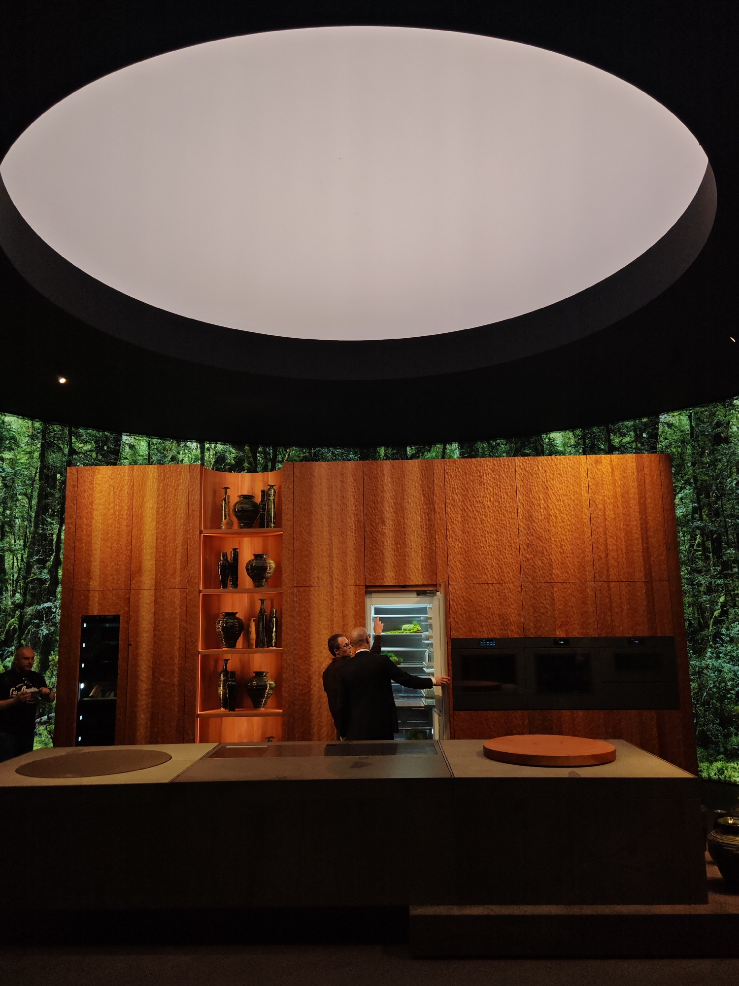 The image captures a modern kitchen display within an indoor setting, likely an exhibition or showroom. The most striking feature is a large, circular, glowing white light fixture dominating the ceiling, set against a dark, curved structure. Below this, a vibrant, large-scale digital screen or mural depicts a dense, green forest with tall trees and moss, serving as a dynamic backdrop to the kitchen.
The central subject is a stylish kitchen unit with extensive wooden cabinetry that has a textured, possibly hammered, finish. Integrated into this wall are several open shelves displaying various black decorative vases or urns, illuminated by warm backlighting. Below these, a built-in refrigerator is open, revealing multiple shelves filled with fresh green produce. Two men in dark suits are positioned near the open refrigerator, one seen from behind reaching inside, and the other partially visible, looking towards its contents.
To the right of the refrigerator, a line of three black integrated kitchen appliances, possibly ovens or microwaves, feature digital displays. One display clearly shows "13:30." In the foreground, a substantial, dark-colored kitchen island or counter is present. It incorporates a circular dark surface on the left and a prominent round, reddish-brown wooden cutting board or trivet on the right.
On the far left of the image, another man in a dark t-shirt, bearing the partial text "Los Angeles," stands looking down at something in his hands. The overall impression is one of a sophisticated, nature-inspired kitchen design showcase, with attendees or presenters interacting with the features.