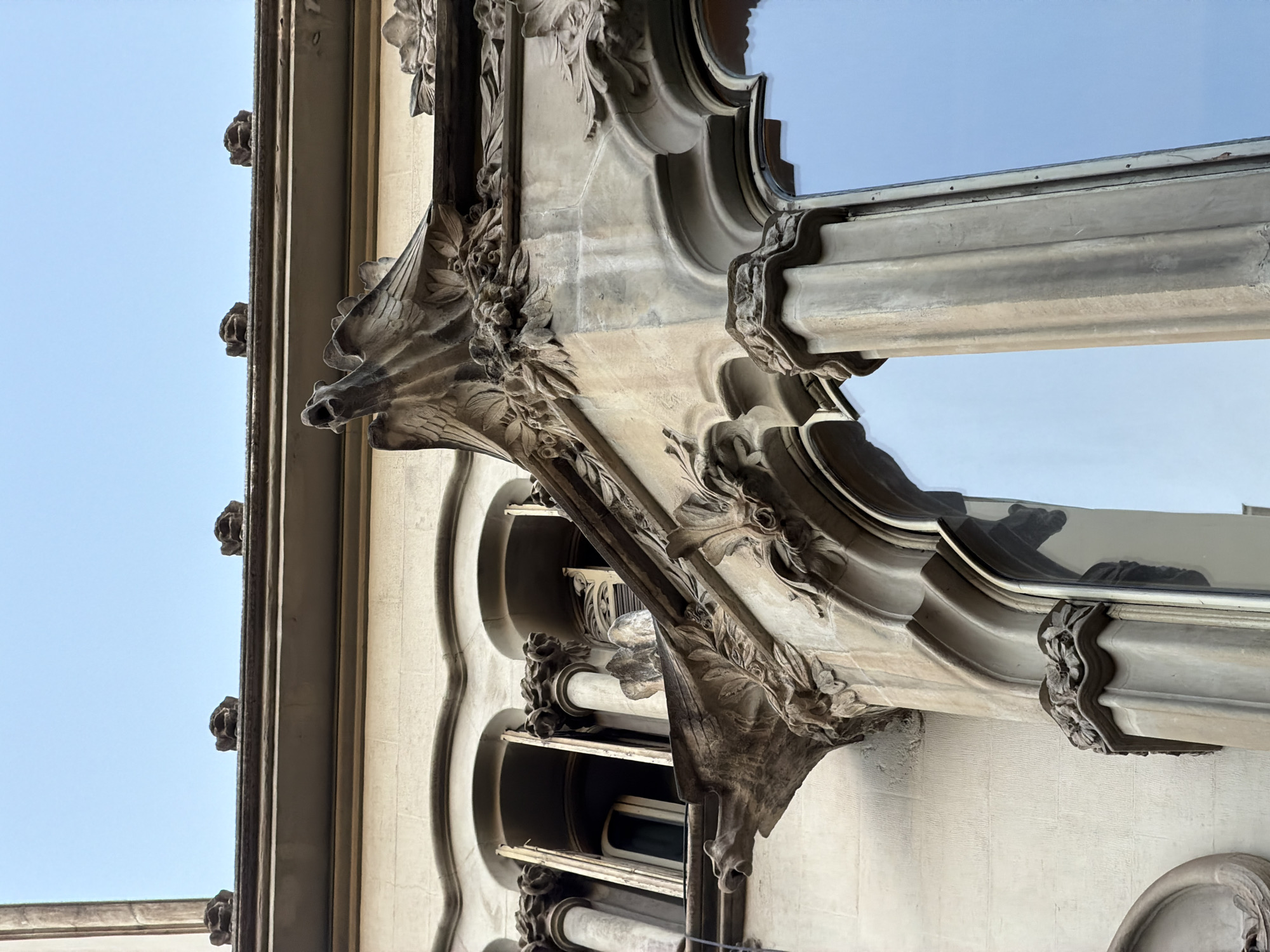 The image displays a close-up, low-angle view of the ornate architectural details of Casa Batlló, located on Passeig de Gràcia in Barcelona, Spain. The building's facade is characterized by its vibrant, undulating forms and decorative elements. In the foreground, a stone balcony features intricate carvings, including what appear to be gargoyles or mythical creatures with open mouths, and floral motifs. The architectural style suggests Art Nouveau, with fluid lines and organic shapes. Behind the balcony, arched windows with segmented openings are visible, framed by decorative stone work and slender columns. The building's exterior is rendered in a light-colored stone, weathered by time. Above the main facade, the rooftop is adorned with small, sculpted figures. The sky above is clear and light blue, reflecting slightly in the large window panes of the building.