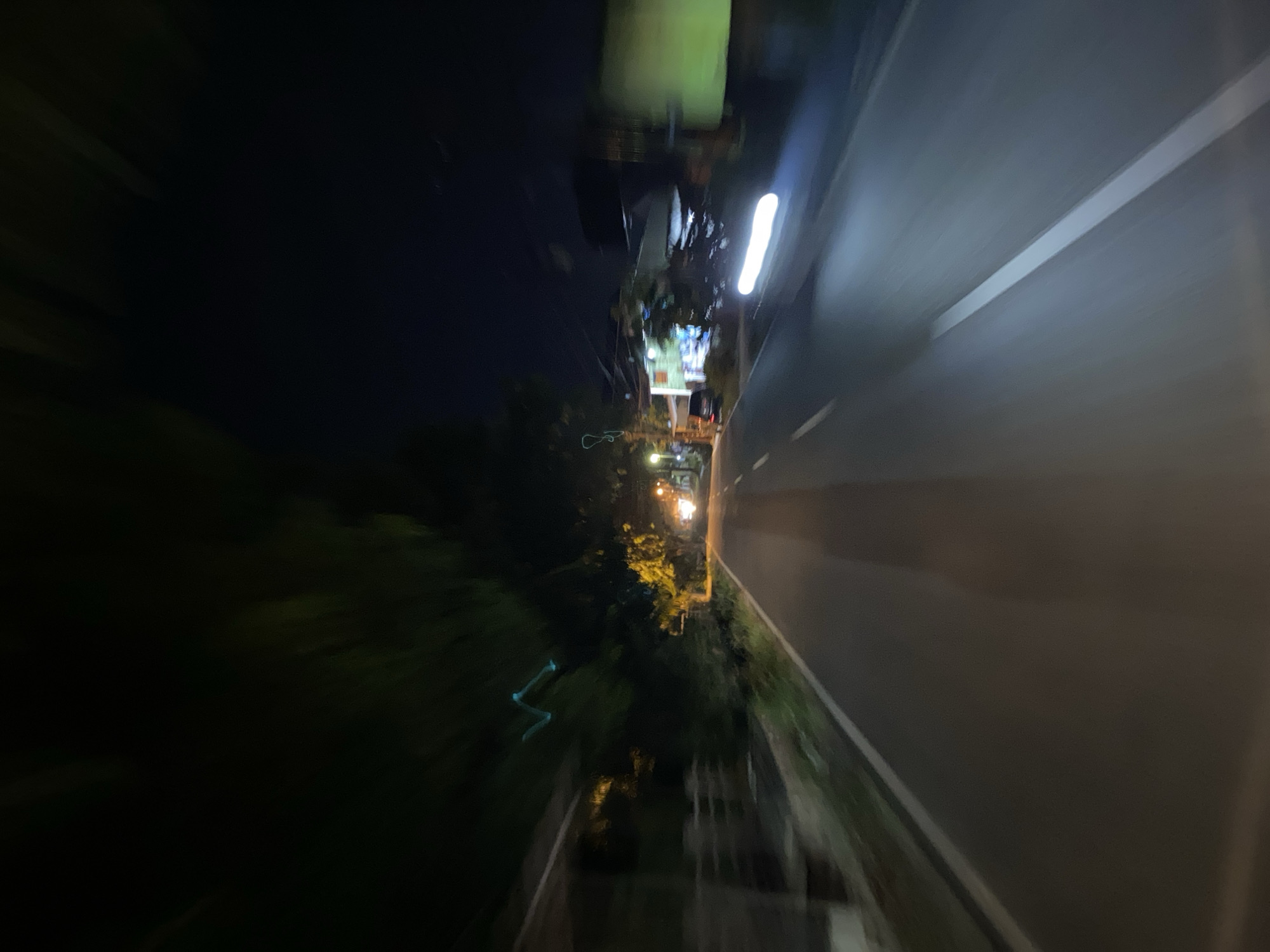 This is a nighttime, motion-blurred photograph of a road in Lhokseumawe, Indonesia. The perspective suggests the image was captured from a moving vehicle. The road is paved and has a white line marking the lane. On the left side of the road, there are trees and some structures that appear to be houses or shops, also blurred by motion. On the right side, there are more trees and buildings, with some illuminated by streetlights or lights from within. In the distance, there are bright lights, possibly from other vehicles or street lamps. The sky is dark, indicating it is night. The overall impression is that of a journey through a suburban or semi-urban area at night.