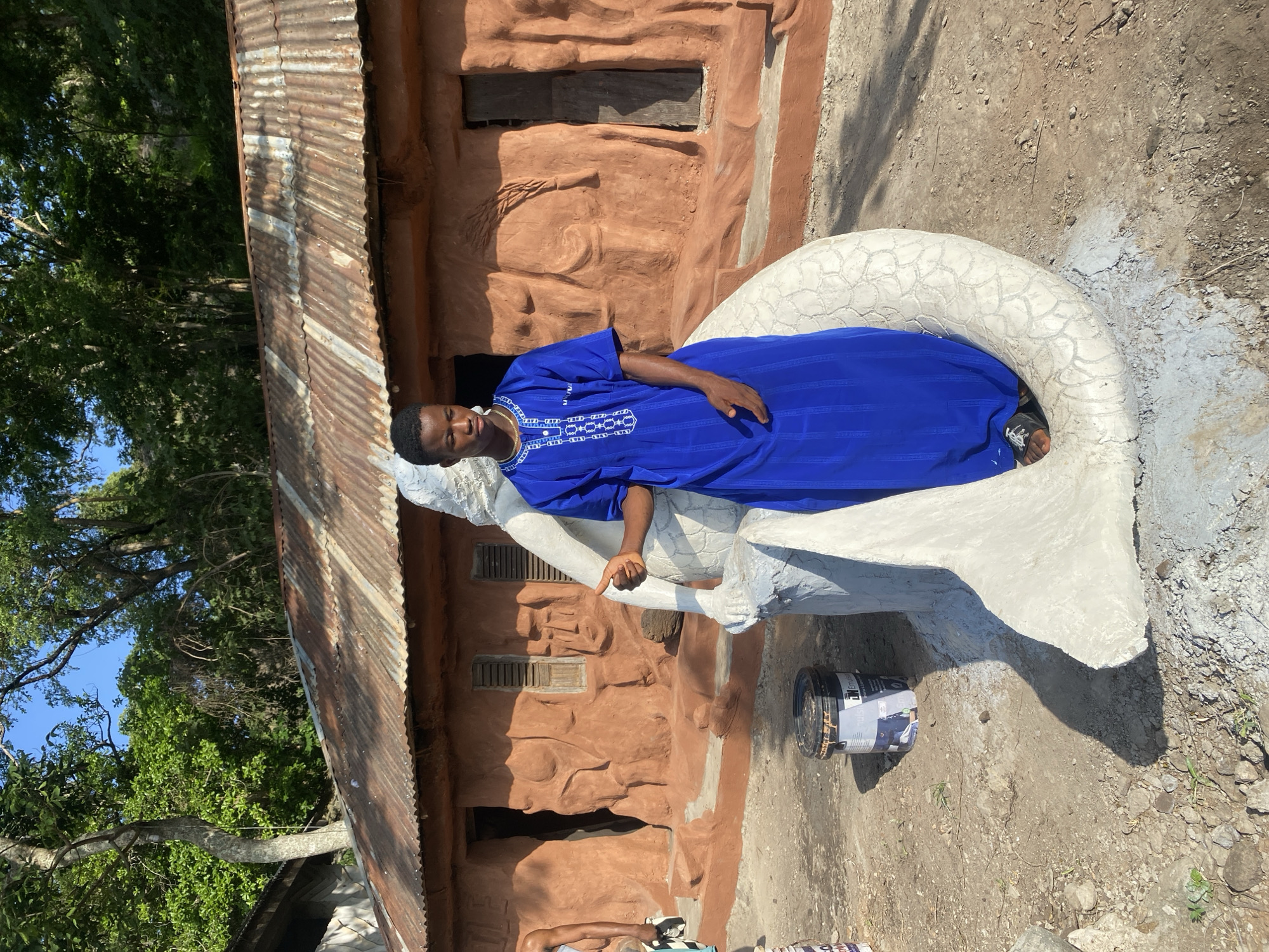 A young Black male stands in front of a building with a rusted corrugated metal roof, in an outdoor environment. He is wearing a blue, ankle-length garment with white embroidered detailing around the neckline and chest. He stands with his left hand resting on a white, serpentine sculpture that appears to be in progress. The sculpture has a scale-like texture. Behind the sculpture and the man, the building is constructed of reddish-brown material and features textured, relief carvings on its facade. Two windows with wooden shutters are visible on the building, as well as an open doorway. To the left of the sculpture, a black bucket with a white label is situated on the dirt ground. The ground is uneven and covered with small rocks and sparse vegetation. Trees with green foliage are visible in the background, casting shadows. The sky above is a clear blue.