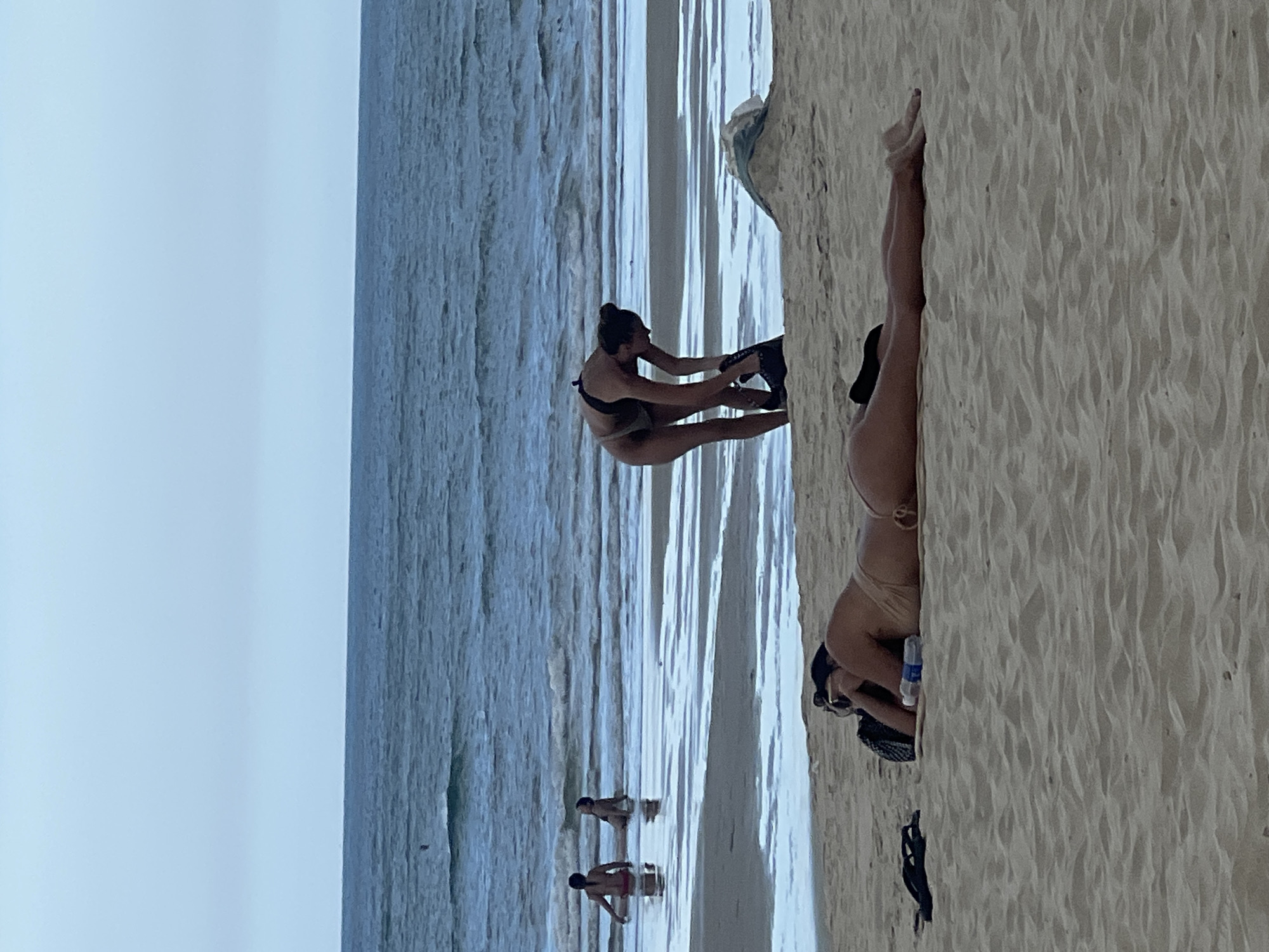 The image depicts a beach scene, likely in Da Nang, Vietnam, under an overcast sky. In the foreground, two individuals are seen on the sandy beach. One person is lying prone on their stomach, wearing a light-colored bikini and sunglasses. Adjacent to them is a black bag, a water bottle, and a pair of flip-flops. Slightly further back, a second person is bending over, partially obscured by their clothing, near the water's edge. In the shallow water further out, two individuals are partially submerged. The ocean waves are visible, and the sky is a uniform pale blue.