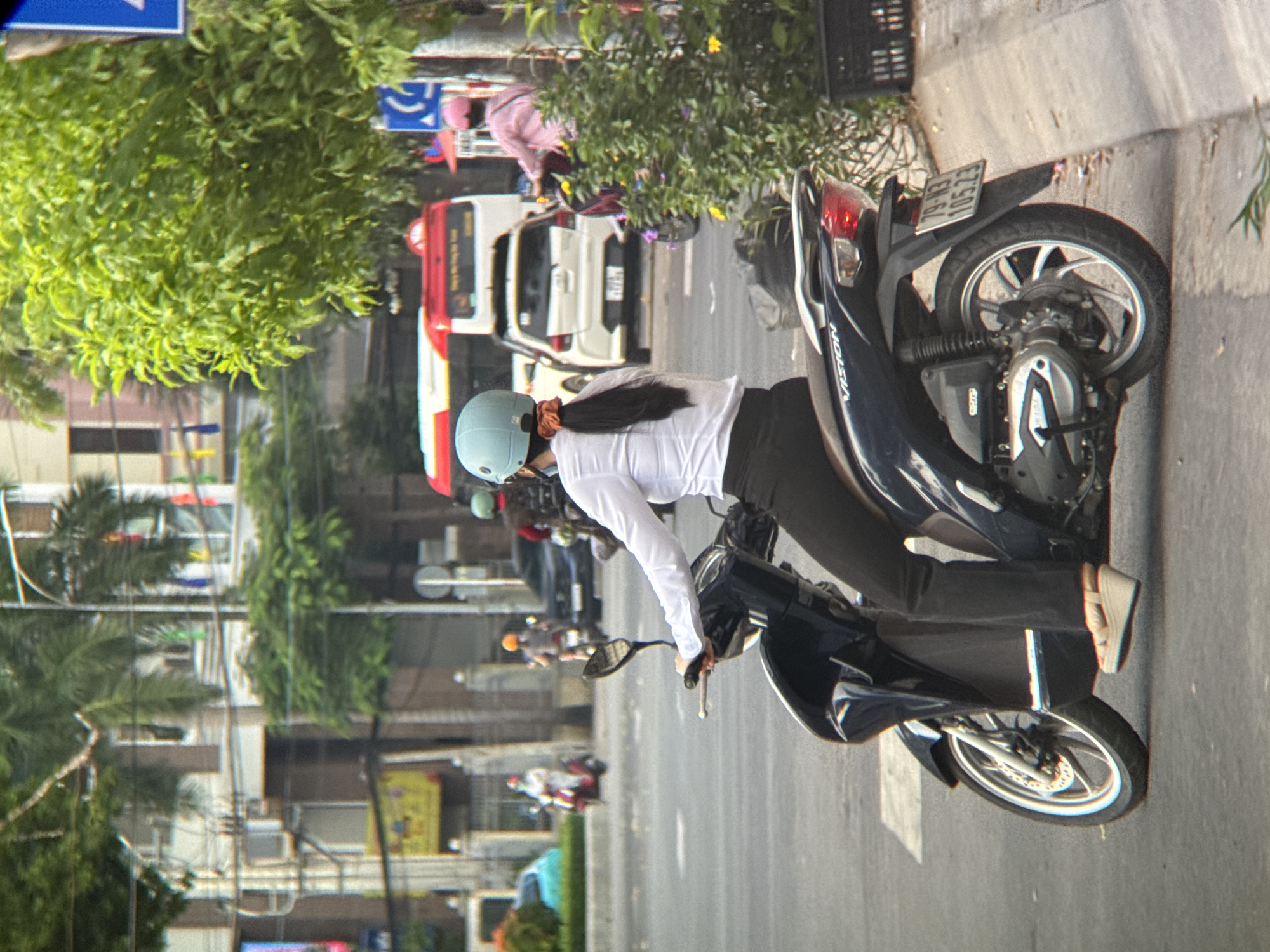 In Nha Trang, Vietnam, a woman wearing a white long-sleeved shirt, black pants, and a light blue helmet rides a dark blue scooter. She has a brown scrunchie in her dark hair, tied back in a ponytail. She is wearing wedge sandals. The scooter's license plate reads "79-M3 105.23". In the background, a red and white bus is visible, along with other vehicles and buildings. Lush green trees line the street. The scene appears to be captured on a daytime street.