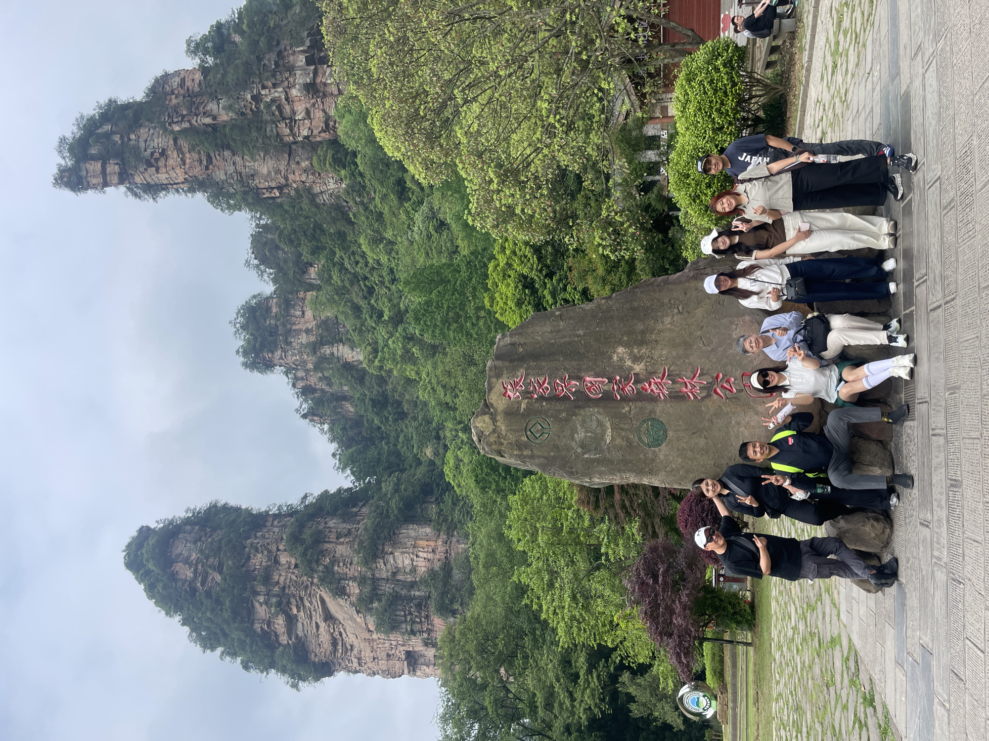 The image shows a group of ten people posing for a photo in front of a large rock with red Chinese characters. The rock is located in a park-like setting in Luoguta, China, with tall, pillar-like rock formations in the background. The landscape is lush with green trees and vegetation. The sky is overcast. The people in the photo are dressed in various casual outfits, and some are making hand gestures. The ground is paved with stone tiles.