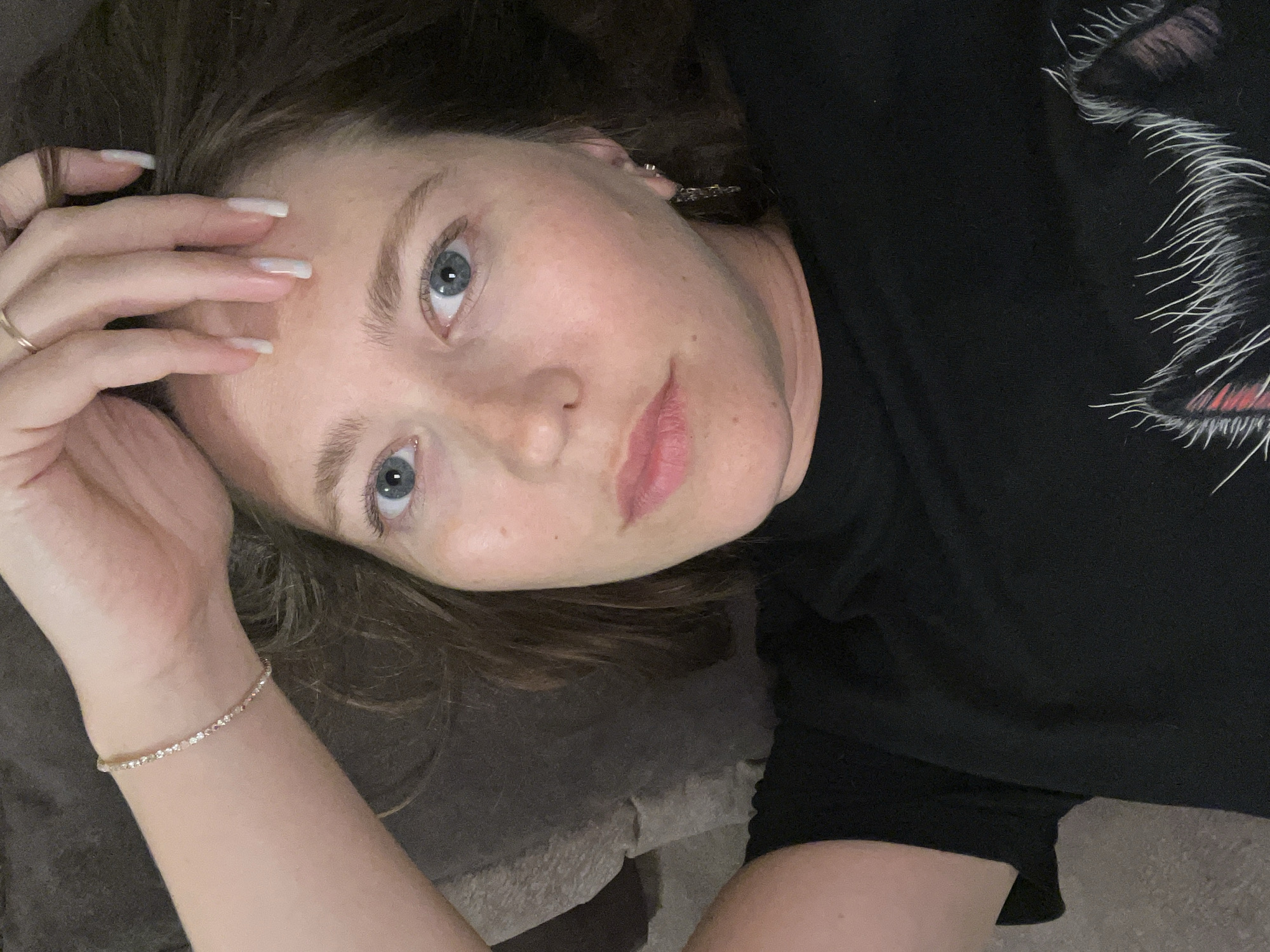 A young woman is captured in a close-up, high-angle shot, lying down and looking directly at the camera. Her head is tilted to the right, and her dark brown, wavy hair frames her face. She has striking light blue eyes with slightly dilated pupils, and her expression is neutral and relaxed. Her lips are lightly colored, possibly with a touch of lip product.
She is wearing a black t-shirt, on which a stylized design is visible on the right side. This design appears to be part of an animal's face, likely a cat, characterized by prominent white whiskers and a hint of red or pink around the eye area. Her left hand is raised and rests near her head, revealing long, well-manicured nails in a light, possibly white or pale pink, color. A simple gold ring adorns her left ring finger, and a delicate, sparkling bracelet is worn on her left wrist. A small, shiny earring can be seen in her right ear.
The setting is indoors, with a dark, textured surface, likely a bed or sofa, serving as the background. The lighting suggests a somewhat dim, indoor environment. The image conveys a casual moment, possibly a selfie or a relaxed portrait. No text is visible in the image. This scene is located in Podgorica, Montenegro.