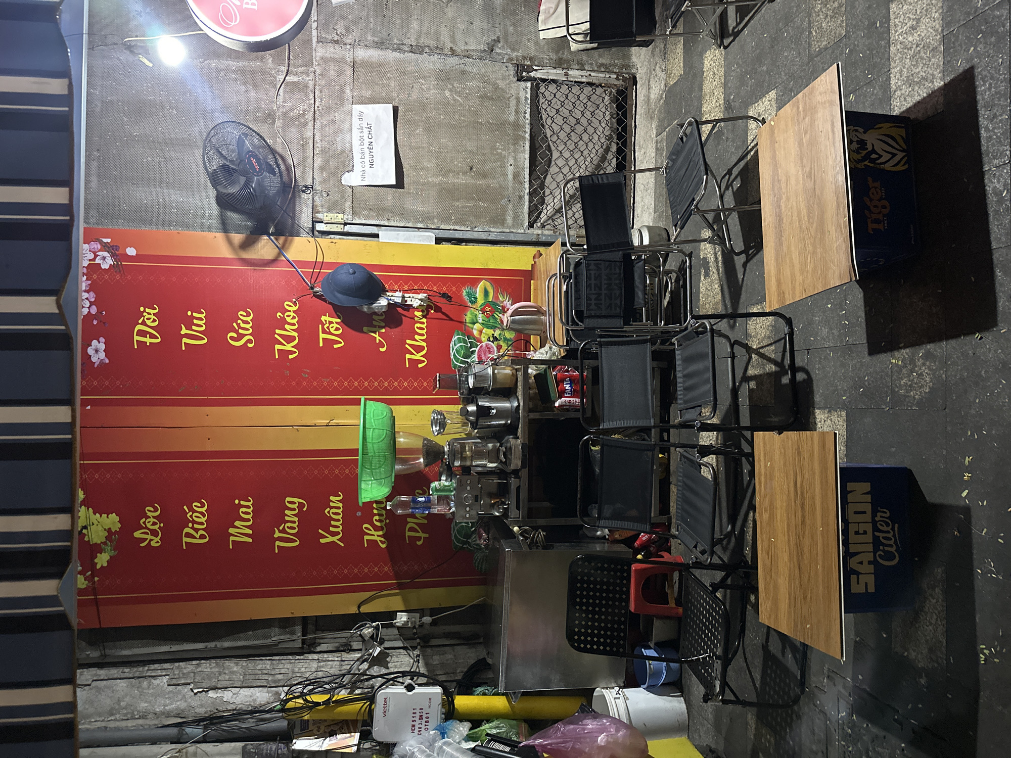 The image depicts a street scene, seemingly an outdoor seating area for a small business, likely a street food vendor or cafe, in Thủ Đức, Vietnam. The environment is urban and utilitarian, suggestive of a densely populated city area.
To the left is a partially visible red and yellow sign, with Vietnamese text in large letters: "Đời Vui Sức Khỏe Tốt An Khang" and then "Lộc Biếc Mai Vàng Xuân Han Phe". Above this sign, there is a cooling fan to the left and the sign "Nhà có bán bột sắn dây NGUYÊN CHẤT" to the right. Tables, each with a wooden top supported by what appears to be stacked beer or soft drink crates, are set up in a row. Metal-framed chairs with black seats and backs are arranged around the tables, indicating an area for customers.
Various items of equipment, which would be expected in the operation of a street food vendor, are also visible. These include food preparation items like a metal countertop, containers, and utensils. Electrical cables and wires run around, which suggests a makeshift setup, typical of street-side businesses.
The ground is paved, with patches of dirt and debris.
The overall impression is one of a functional, informal business operating in a public space. There are no people visible in the shot, so the scene appears to be at a moment when the business is either not open or not busy.