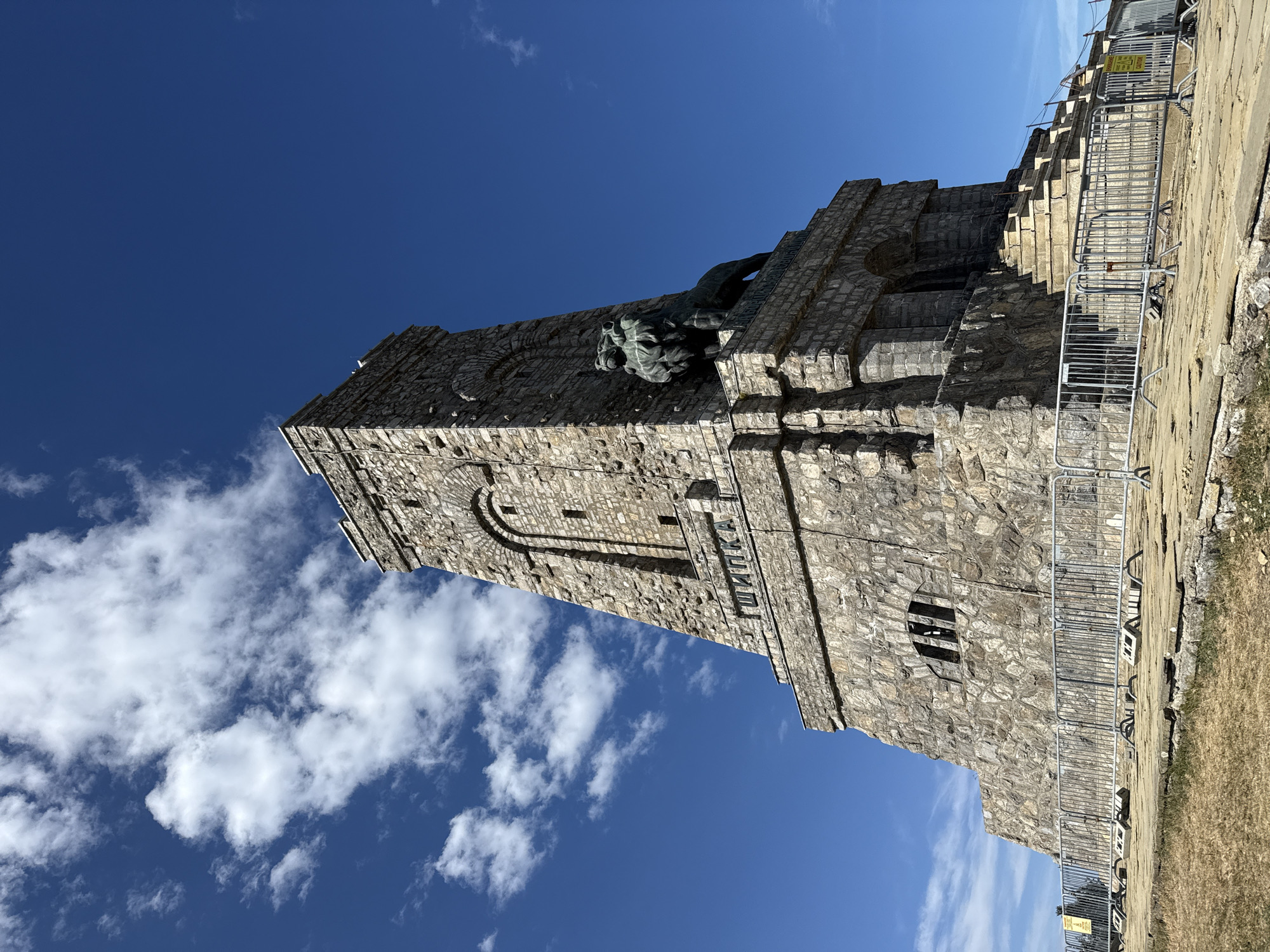 This is a low-angle, eye-level, outdoor, daytime photograph of the Monument to the Freedom of Bulgaria, also known as the Shipka Monument, located on Stoletov Peak near Shipka, Bulgaria. The monument is a massive stone structure with a tall, slender tower that tapers towards the top. It is adorned with arched windows and a large, dark bronze statue of a lion sitting on a pedestal on its right side. There are Cyrillic inscriptions on the monument, including "ШИПКА" on a band below the lion statue. The monument is surrounded by a metal barricade, and the ground in front of it is paved with stone slabs, some of which appear cracked and uneven. Dry, sparse vegetation covers the ground in the foreground. The sky is a vibrant, clear blue, with scattered white, fluffy clouds. The scene is quiet and appears to be unoccupied by any individuals.