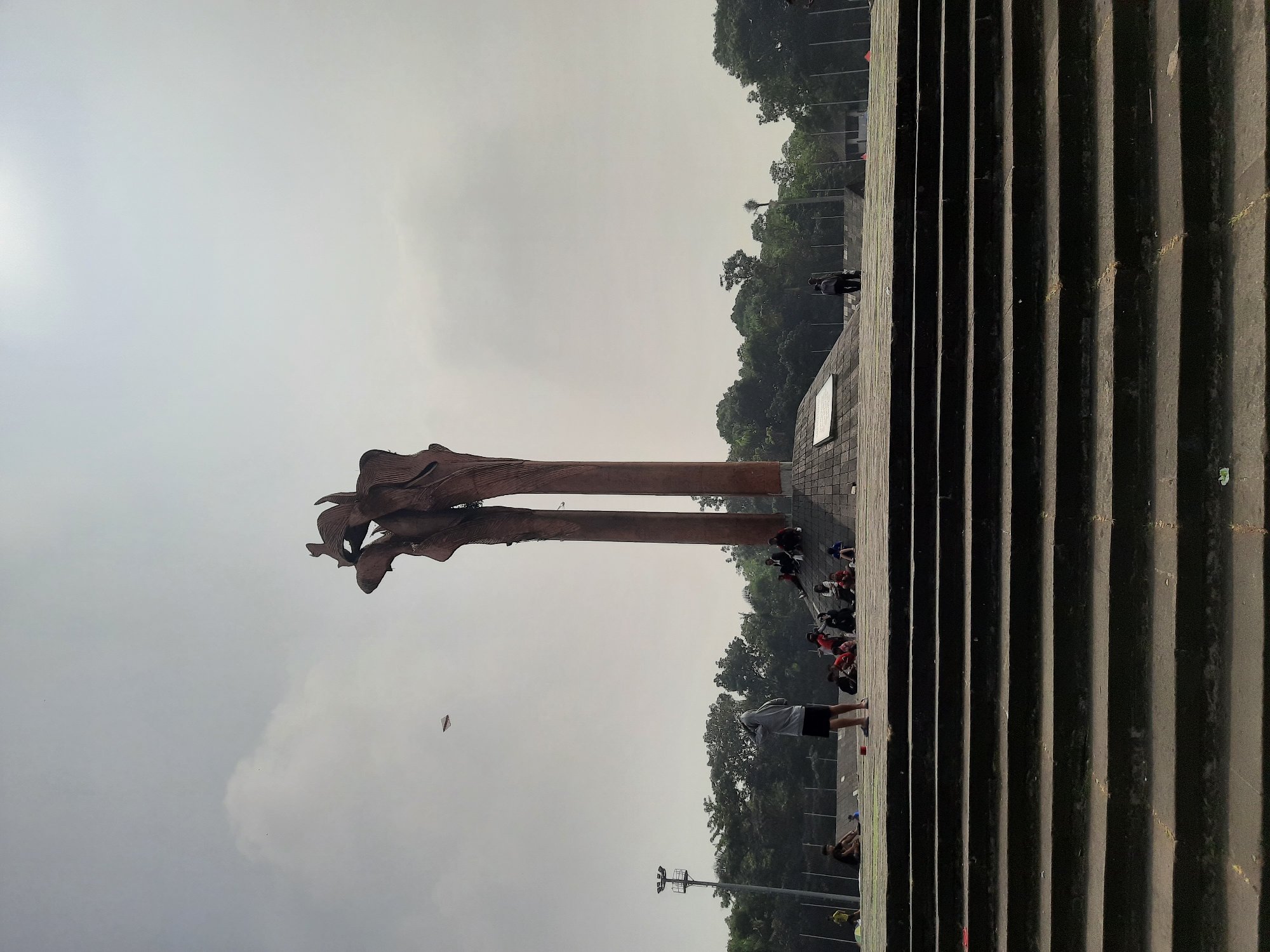 The image depicts the Monumen Perjuangan Rakyat Jawa Barat, also known as the Sate Monument, located in Bandung City, Indonesia. The monument, a tall, abstract sculpture resembling flames or reaching hands, dominates the center of the frame. It is made of weathered metal and stands on a raised platform. In the foreground, a wide set of stone stairs leads up to the monument. A group of people are scattered on the stairs and the platform, some sitting and others standing. One person in the mid-ground, wearing a grey hoodie and shorts, is facing away from the camera, seemingly observing something in the sky. Another individual in the background is flying a kite. The environment is characterized by a hazy, overcast sky, with lush green trees forming a backdrop behind the monument and the surrounding area. The overall atmosphere suggests a public gathering or recreational space on a cloudy day.