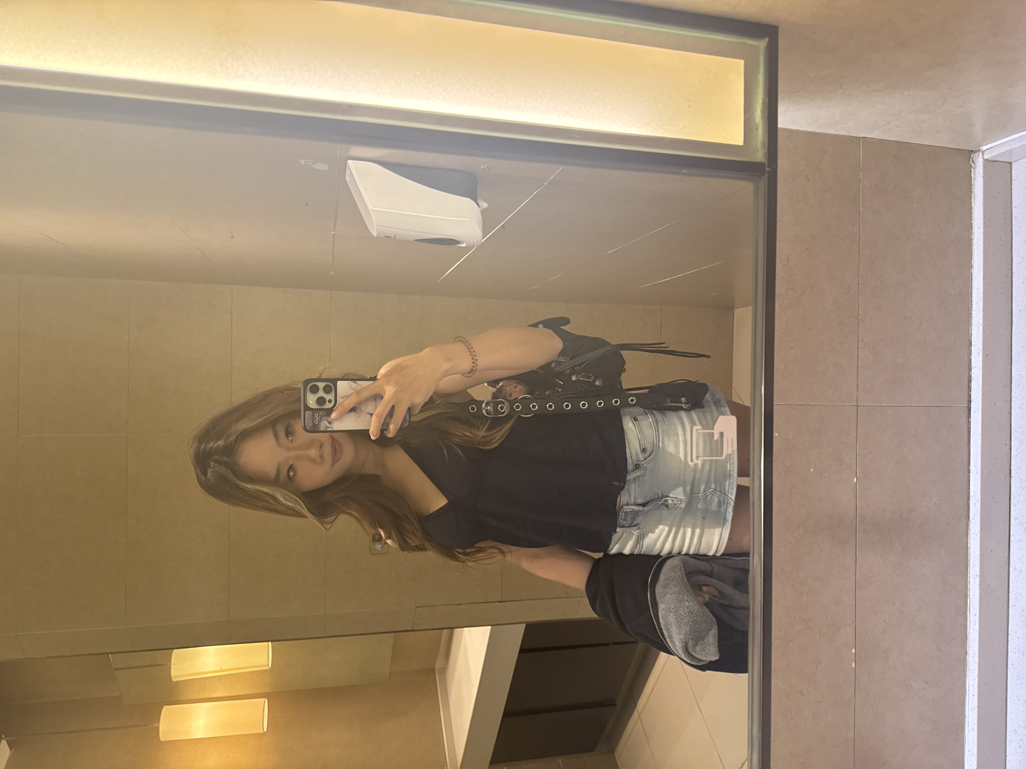 A young woman is taking a selfie in a public restroom. She is holding a smartphone in her right hand. The woman is wearing a black t-shirt, light blue denim shorts, and a studded belt. A gray jacket is slung over her shoulder. The restroom walls and fixtures are light brown. A white dispenser is visible on the wall behind the woman. The image appears to be a reflection in a mirror. It is impossible to confirm that this image was taken in Yuzhong District, China, based on the image alone.