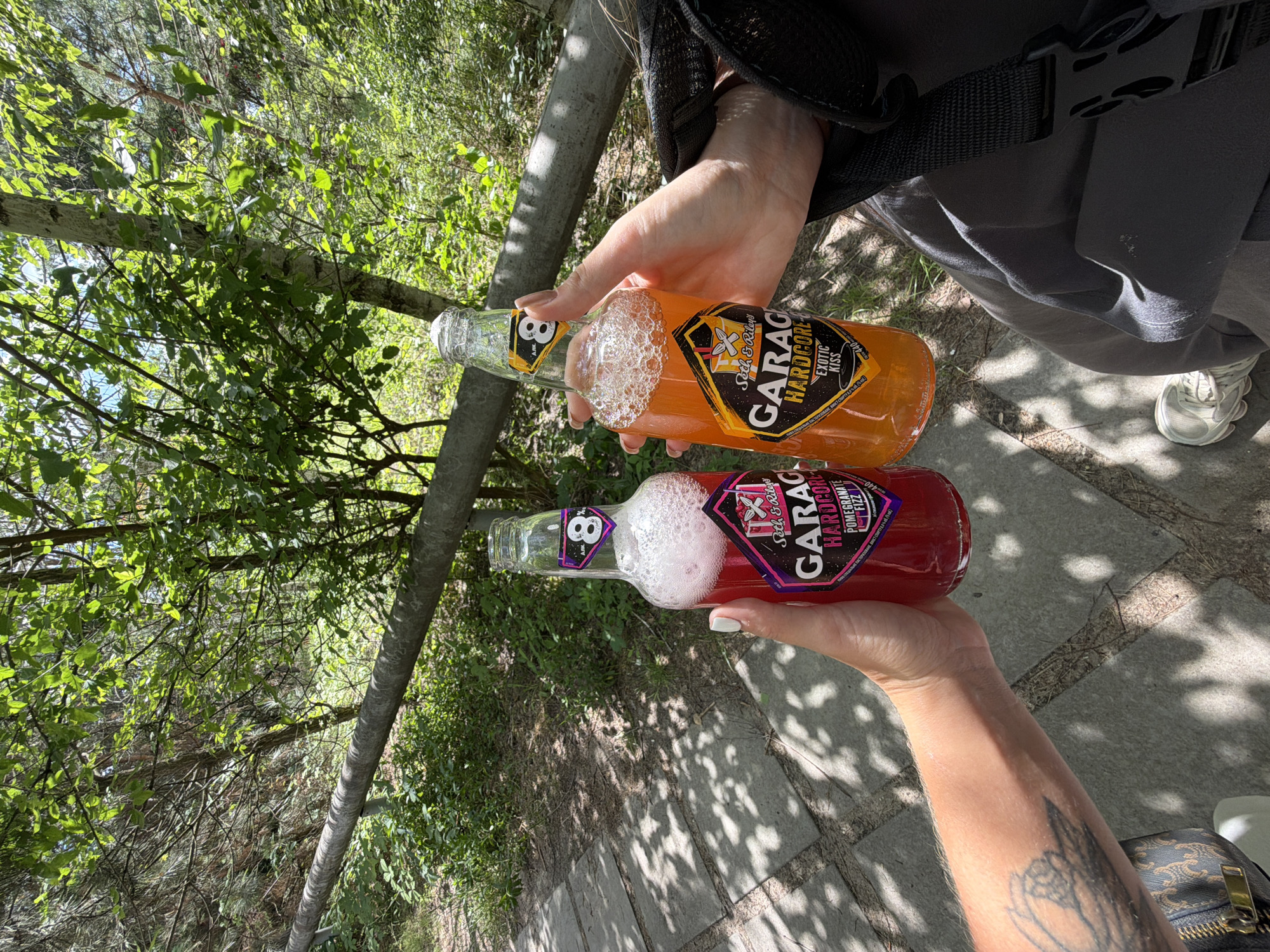 Two people are toasting with bottles of Garage Hardcore beverages in a park. The person on the left holds a bottle of Pomegranate Fizz, which is a deep red color with a foamy head. The person on the right holds a bottle of Exotic Kiss, which is an orange color with a foamy head. Both bottles have the Garage Hardcore logo and an "8" in a yellow triangle. The background shows lush greenery with trees and a dirt path. This is likely in the city of Obukhivka, Ukraine, given the context.