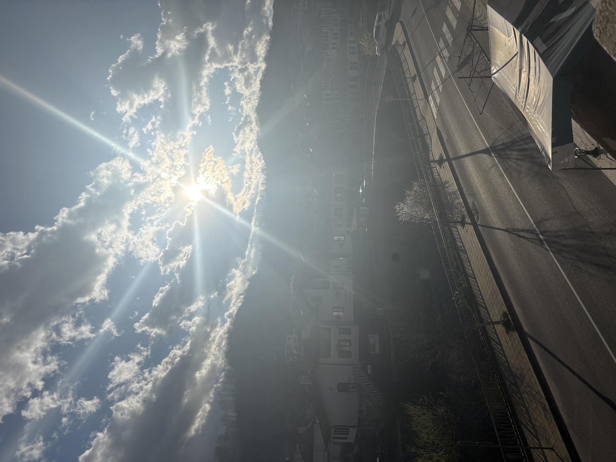 The image shows a street scene in Zlatograd, Bulgaria, viewed from an elevated position. The sun shines brightly through scattered clouds. A road runs along the right side, with streetlights and fencing along the sidewalk. Buildings are visible in the middle ground, partially obscured by haze. A crosswalk marks the intersection in the foreground.