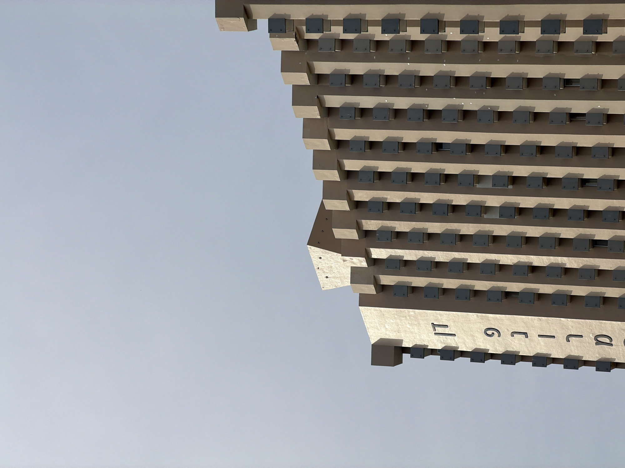 The image displays the upper section of the Dan Panorama Hotel in Tel Aviv, Israel, viewed from an upward angle against a clear, light blue-grey sky. The building's facade is constructed of light beige material, featuring a prominent and repetitive architectural pattern. This pattern consists of numerous dark grey, square-shaped projecting elements, likely balconies or sunshades, arranged uniformly in vertical columns. These projections cast distinct shadows to their left, indicating a light source from the right. On the vertical left side of the building, large, dark grey Hebrew letters are arranged vertically, spelling "דן פנורמה" (Dan Panorama). The building's roofline culminates in a stepped, castellated design composed of cuboid structures. No individuals or additional objects are present in the frame.