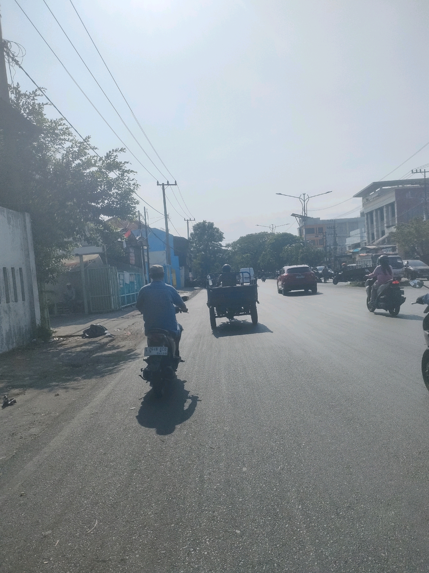 A street scene in Surabaya, Indonesia, on a bright, possibly hazy, day. The shot is taken from the perspective of someone riding a motorcycle, looking forward.
In the foreground, a person in a blue checkered shirt is riding a dark-colored motorcycle. The motorcycle has a license plate that reads "2648 GCO 04-20". To the left of the road, there is a white building with vertical lines and a green fence. Trees with dense foliage are visible behind the fence. Some debris is scattered on the ground near the fence.
The road itself is paved and appears to be a multi-lane street. Several other vehicles are present. Further ahead on the road, a three-wheeled cargo motorcycle is being ridden by a person wearing a helmet. To the right, a woman in a pink shirt and helmet is riding a motorcycle, with a passenger behind her. A red SUV is also visible in the middle distance, along with other vehicles that are less distinct.
In the background, buildings of various sizes and styles are lined up along the street, suggesting a commercial or mixed-use area. Power lines and streetlights are visible overhead, strung between utility poles. The sky is bright and washed out, indicating the sun is high and possibly obscured by haze. The overall impression is a busy urban street with ongoing traffic.