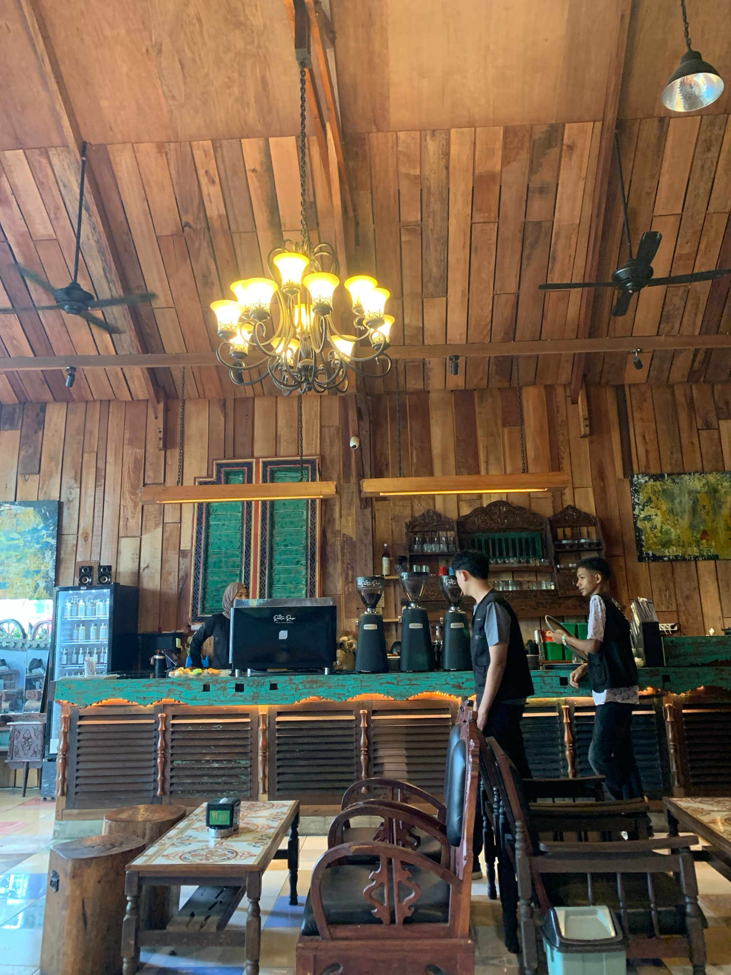 This is a medium shot taken from a slightly low angle, looking up into a coffee shop with a prominent wooden ceiling and walls, featuring a rustic aesthetic. Two male baristas, dressed in black vests over their shirts, are visible behind a turquoise counter. One barista, on the left, is facing away from the camera and appears to be working with equipment, possibly making coffee. The other barista, on the right, is looking towards the left and holding a round object, possibly a tray or a plate. A female barista, wearing a hijab and a black shirt, is partially visible on the left side of the counter, also engaged in work.
The counter area is equipped with multiple coffee grinders and a large black coffee machine. Behind the baristas, there are shelves filled with glasses and bottles, and a decorative wooden structure hangs above them. To the left of the counter, a refrigerator is visible, displaying various bottles.
In the foreground, several wooden tables and chairs are arranged. A small table in the center has a decorative tiled top and a tissue box. Large wooden tree stumps serve as additional seating or side tables. Two ceiling fans are mounted on the wooden ceiling, and a large, ornate chandelier hangs above the counter. Paintings with abstract designs are displayed on the walls. The floor has a checkered tile pattern. The overall ambiance suggests a cozy and somewhat vintage coffee shop environment. The image was taken in the city of Lhokseumawe, Indonesia, likely within a coffee shop named "Retta Popo," as indicated by branding on the coffee machine.