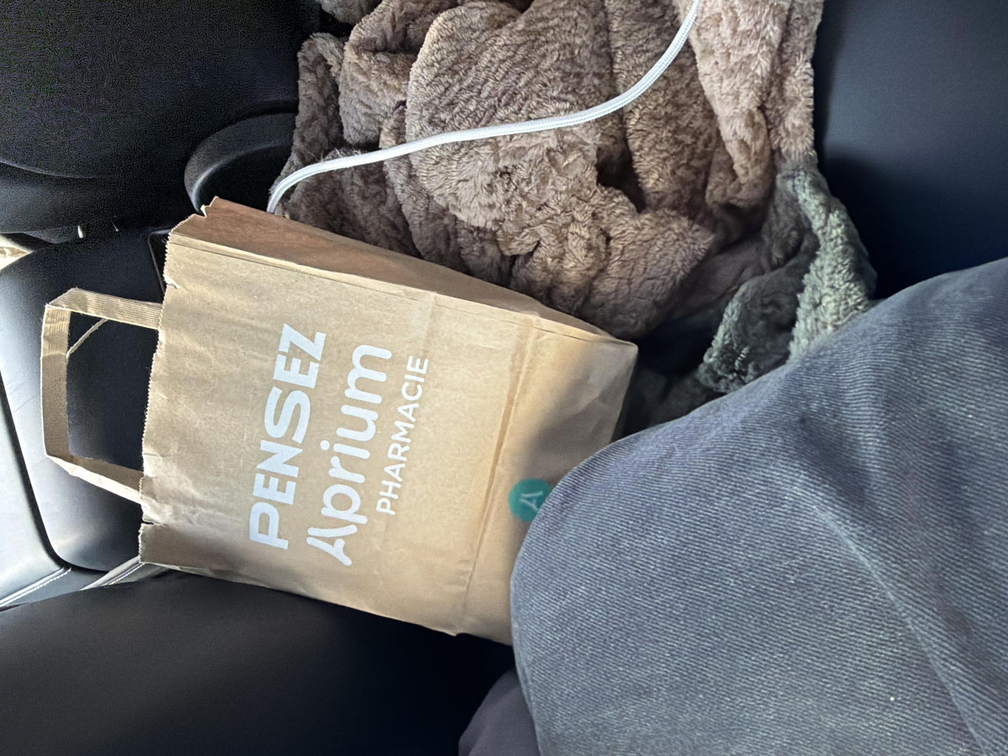 A brown paper bag with the branding "PENSEZ Aprium PHARMACIE" is placed on a car seat. A person's leg, clad in blue denim jeans, is visible in the foreground. A plush, brown blanket is partially covering the bag and the seat. A white cable is also visible, running across the blanket. A small, teal-colored circular sticker with the letter "A" is affixed to the paper bag. The image appears to have been taken inside a vehicle. The location is Ödsmål, Sweden.