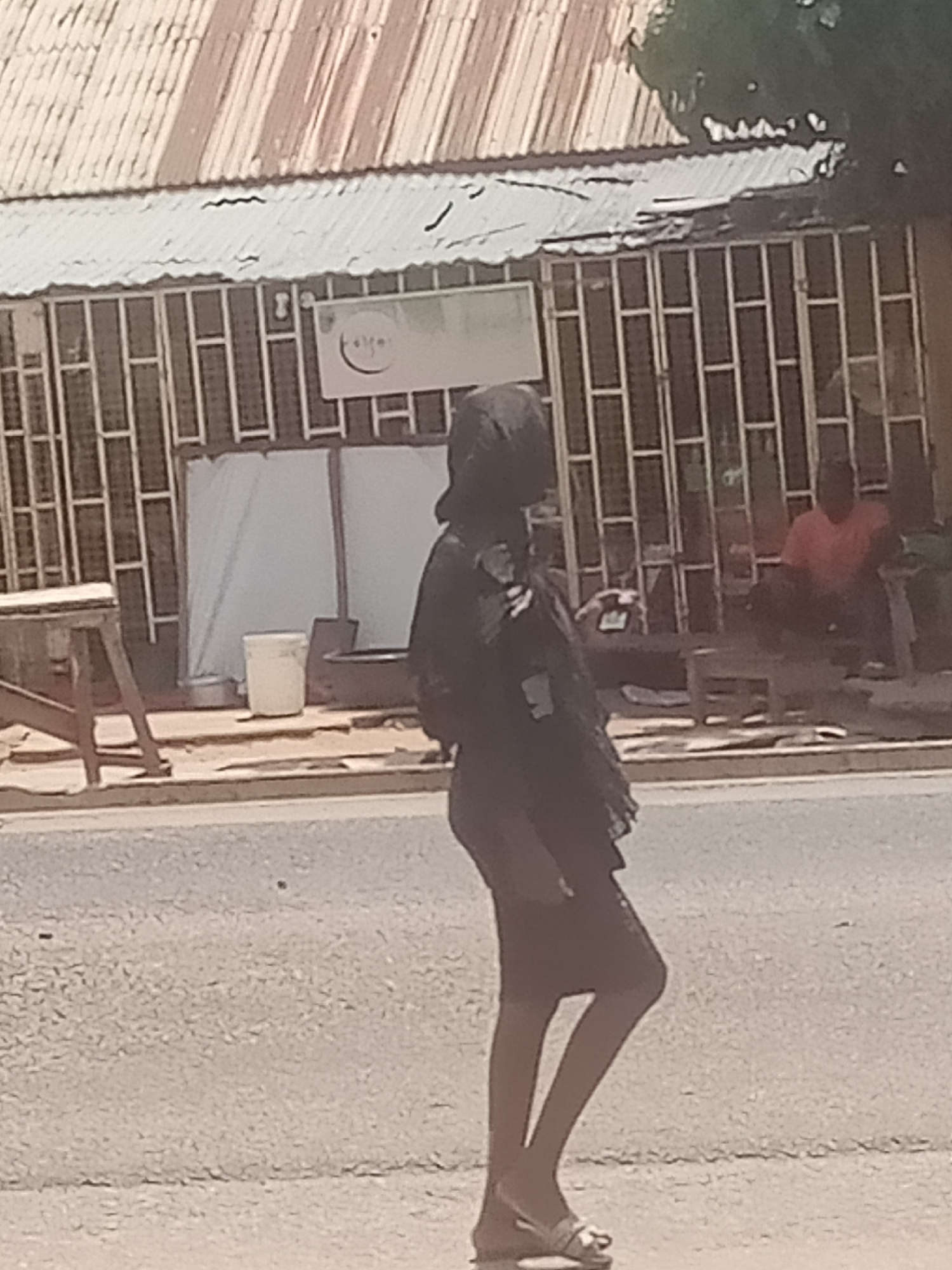 A woman wearing a dark headscarf and dark clothing walks on a paved road in Jalingo, Nigeria. She is seen from the side, walking from left to right across the frame. In the background, there is a building with a corrugated metal roof and bars on its windows. A white bucket is visible near the building, as well as a person in a red shirt sitting on a bench. The overall scene appears to be a street in a commercial or residential area.