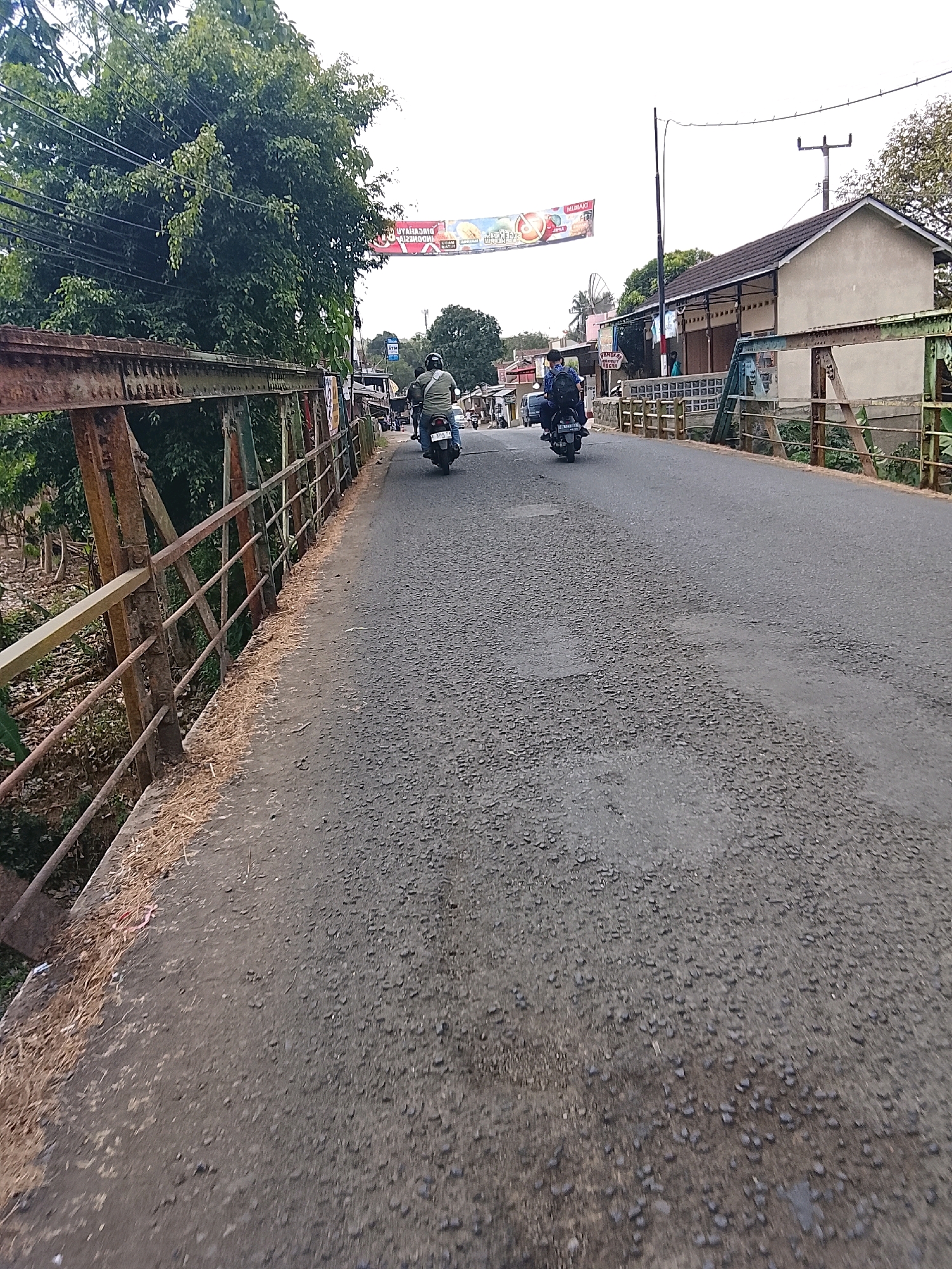 The image shows a street scene in Pamarican, Indonesia, likely on a bridge. Two individuals are riding motorcycles on a slightly deteriorated asphalt road. The motorcycle on the left is ridden by a person wearing a green shirt and a helmet, while the motorcycle on the right is ridden by a person wearing a blue shirt and a backpack. To the left of the road, there is a rusty metal railing with a weathered wooden or metal beam on top, bordering what appears to be a sloped embankment with vegetation and a small stream. On the right side of the road, there are buildings with tiled roofs and visible storefronts, some with awnings. A banner with Indonesian text and images of fruit hangs overhead. Utility poles and wires are also visible. The sky is overcast.