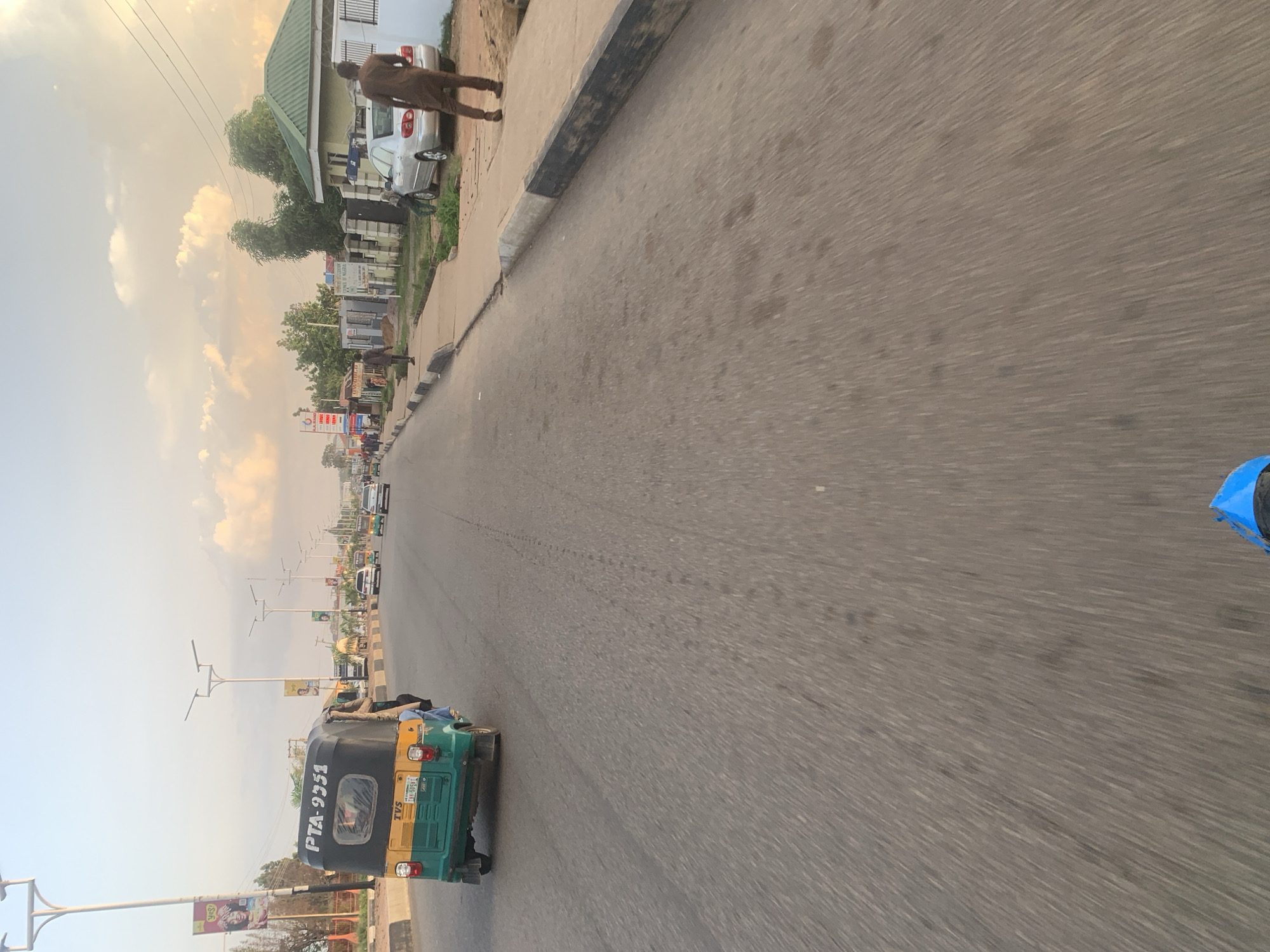 The image depicts a street scene in Jos, Nigeria, likely taken from a moving vehicle. A green and yellow auto-rickshaw, with the license plate PTA-9351, is in the left foreground, its passenger area partially occupied. The road is paved, with a distinct curb on the right side, separating it from a sidewalk. Beyond the sidewalk, there are buildings and vegetation. A man in a brown traditional outfit is walking on the sidewalk, and a silver sedan is parked nearby. Other vehicles and streetlights are visible further down the road, suggesting an urban environment. The sky is partly cloudy, with soft, warm light indicating it is likely late afternoon or early evening.