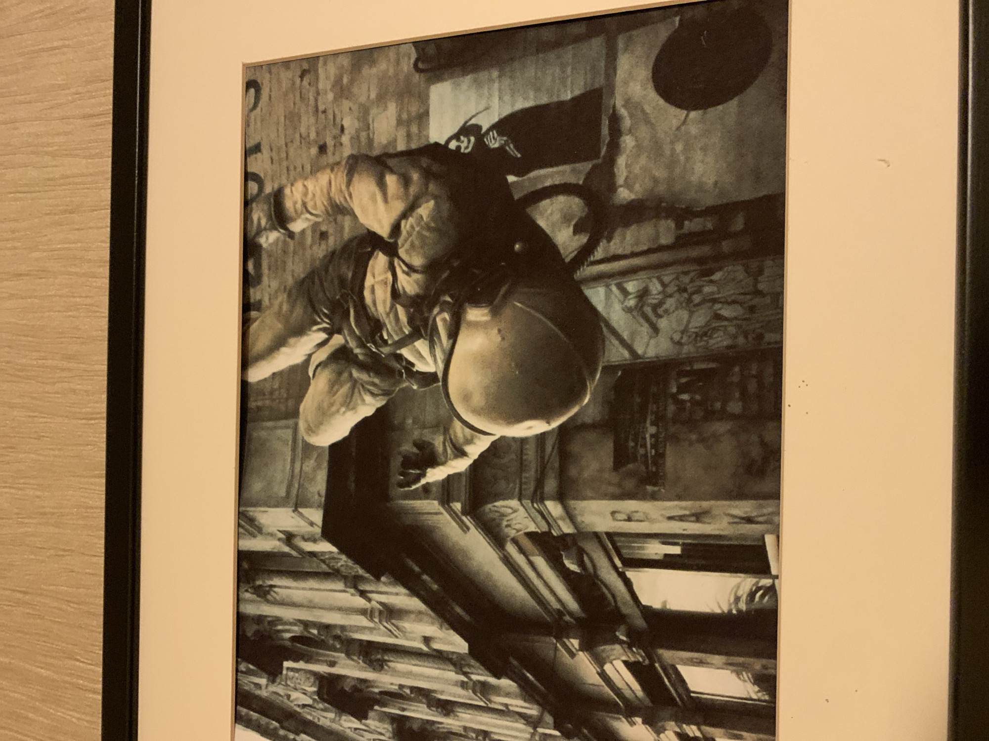 The image shows a black and white photograph of a person in mid-air, suspended by a rope or wire, with a building in the background. The person appears to be wearing a light-colored jumpsuit and a helmet, and is positioned as if they are falling or swinging. The building is old and ornate, with details like columns and windows. There is some graffiti on the wall. The scene is dynamic and suggests an urban environment.