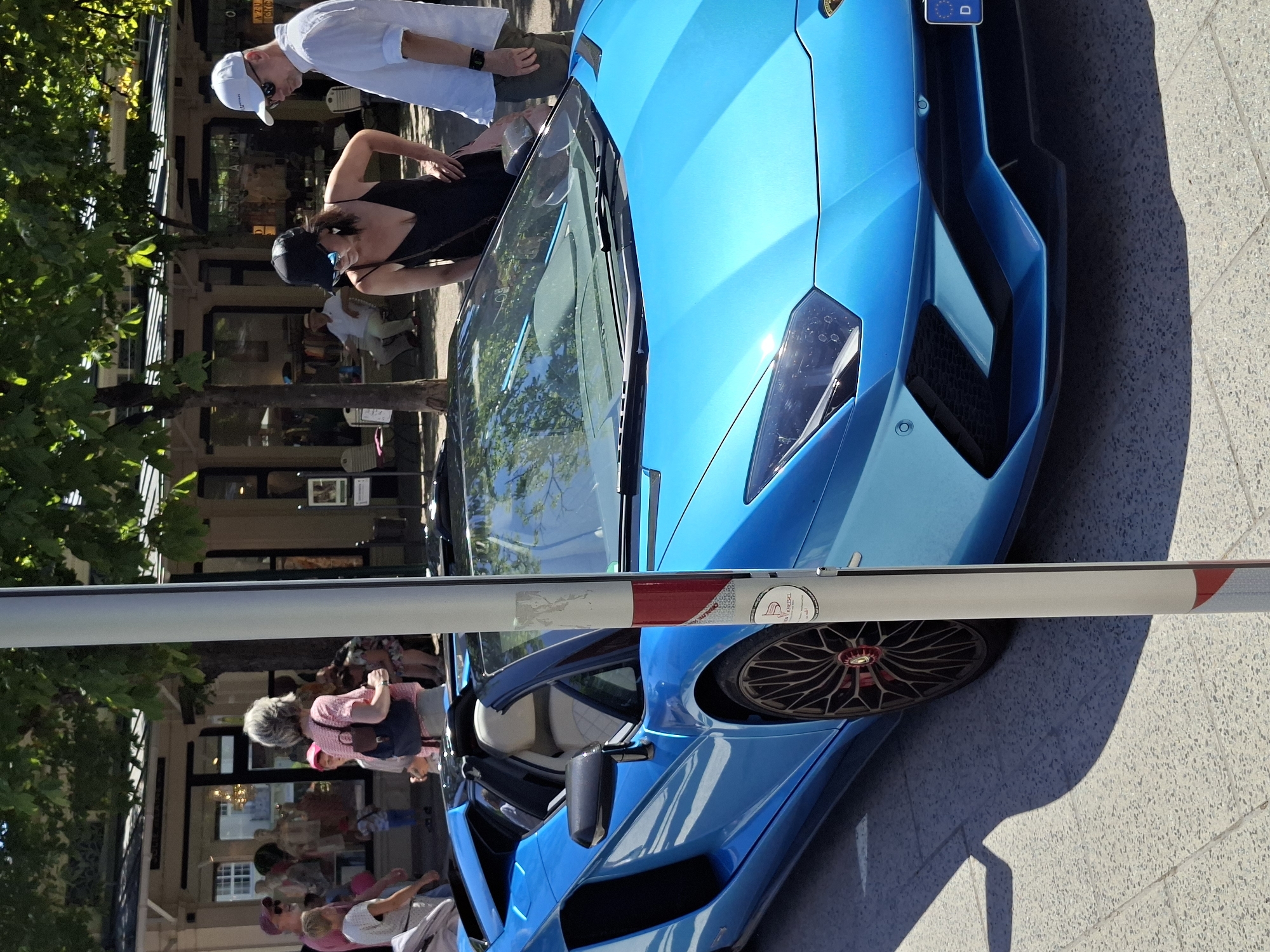A bright blue Lamborghini Aventador convertible is parked on a pedestrian street in Baden-Baden, Germany, possibly in front of the shops lining the Lichtentaler Allee. The car's black convertible top is down, revealing its cream-colored interior. In the background, several people are visible browsing storefronts or walking along the street. Two individuals, a woman in a black dress and a man in a white shirt and cap, are standing near the car, appearing to admire it. Another group of people, including a child in a pink hat, are further in the background near a shop entrance. The street is paved with light-colored stones, and trees with green foliage provide shade. A vertical white pole with red and blue accents stands in the foreground, partially obscuring the view of the car. The license plate on the car is partially visible, showing a German "D" plate.