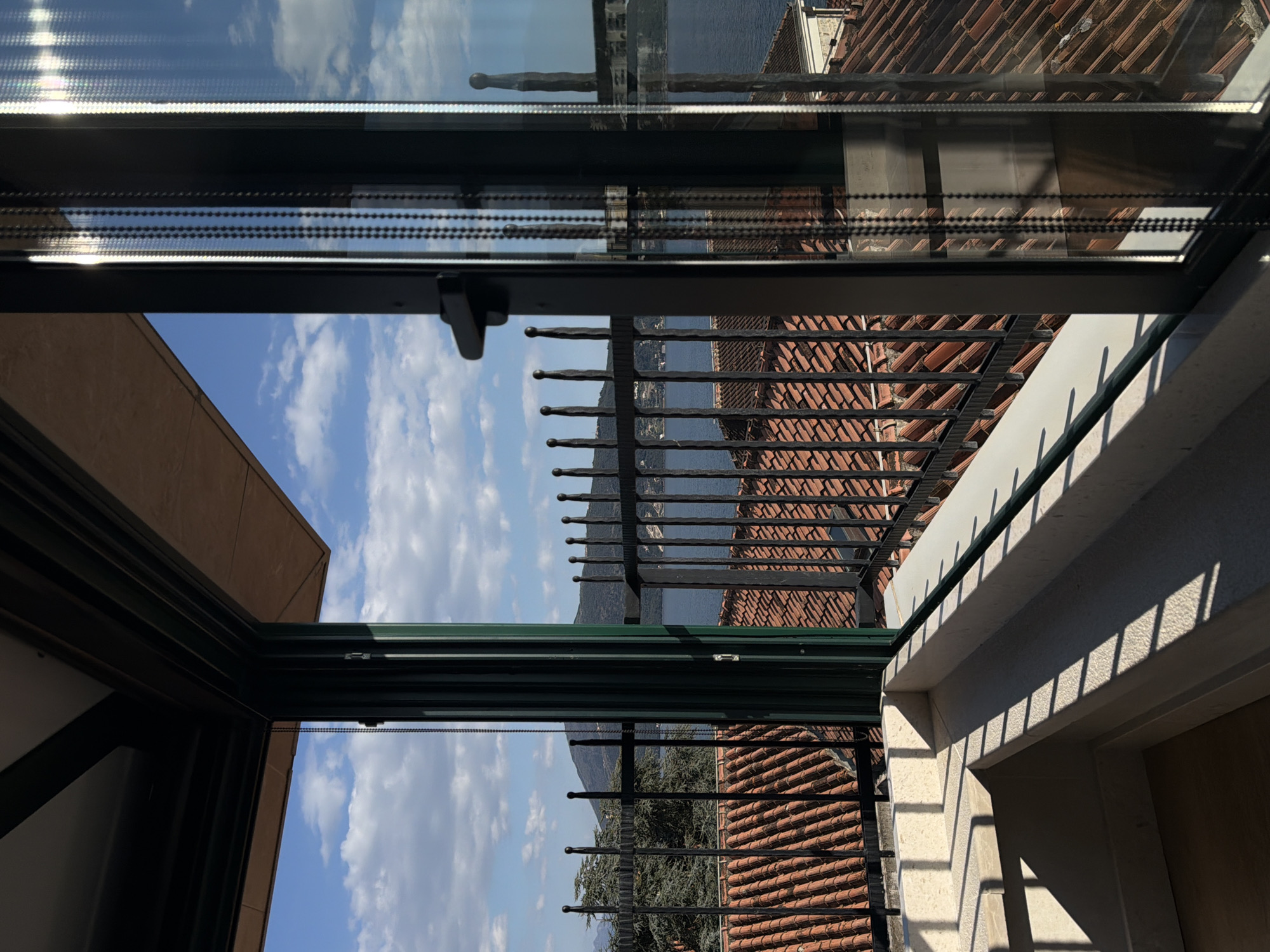 The image presents a view of a landscape through what appears to be an open window or balcony enclosure. The primary focal points are:
1. **Window/Balcony Structure:** A dark-colored metal frame divides the view. A chain-like cord hangs near the top frame, suggesting a window shade or blind mechanism. The frame extends both horizontally and vertically, creating a sense of depth.
2. **Rooftops and Landscape:** Beyond the frame, tiled rooftops are visible, likely belonging to nearby buildings. Further in the background, there is a body of water, possibly a bay or lake, with landmasses across it. The sky above is blue with scattered clouds.
3. **Metal Railing:** A wrought-iron-style metal railing runs along the balcony.
Based on the provided location of Trebesin, Montenegro, it's plausible the body of water is part of the Bay of Kotor.