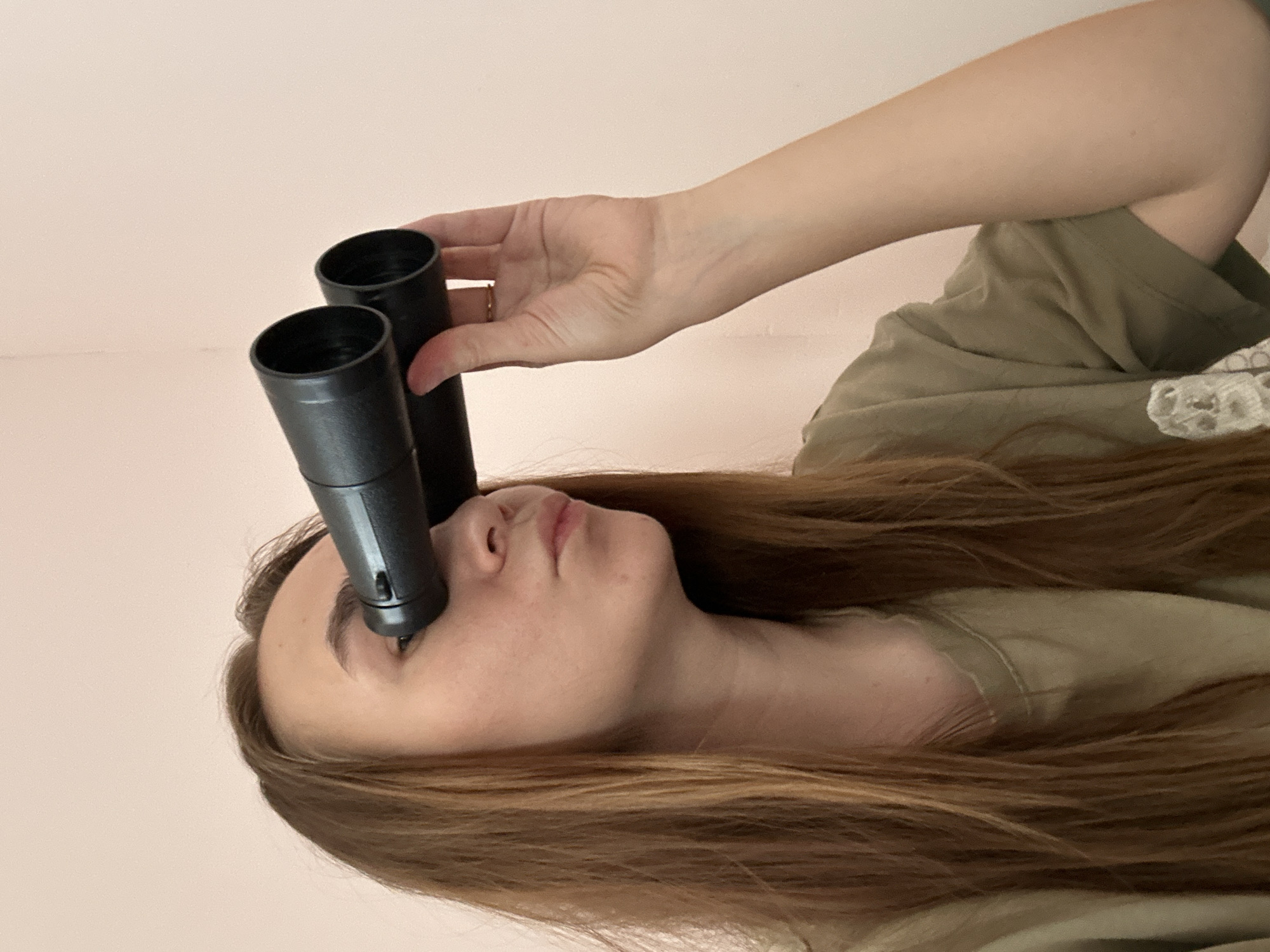 In an indoor setting, likely in Kharnavy, Russia, a young woman with long, flowing brown hair is depicted. She is positioned against a plain, light pink background. The woman wears an olive-green garment, possibly a shirt or dress, which features white lace detailing on the shoulder area.
Her right arm is extended, and her hand holds two black, cylindrical objects, which appear to be hollow tubes or parts of a larger device. One of these cylinders is placed directly over her right eye, while the other is positioned next to it, potentially covering her left eye or resting near it. Her head is tilted slightly back, and she appears to be looking forward or upward, with a neutral, perhaps contemplative or curious, expression. A subtle gold ring can be seen on one of her fingers.
The image is rotated 90 degrees counter-clockwise, presenting the subject horizontally. There is no visible text in the scene. The overall impression is one of a posed moment, possibly exploring an unusual way of seeing or interacting with objects.