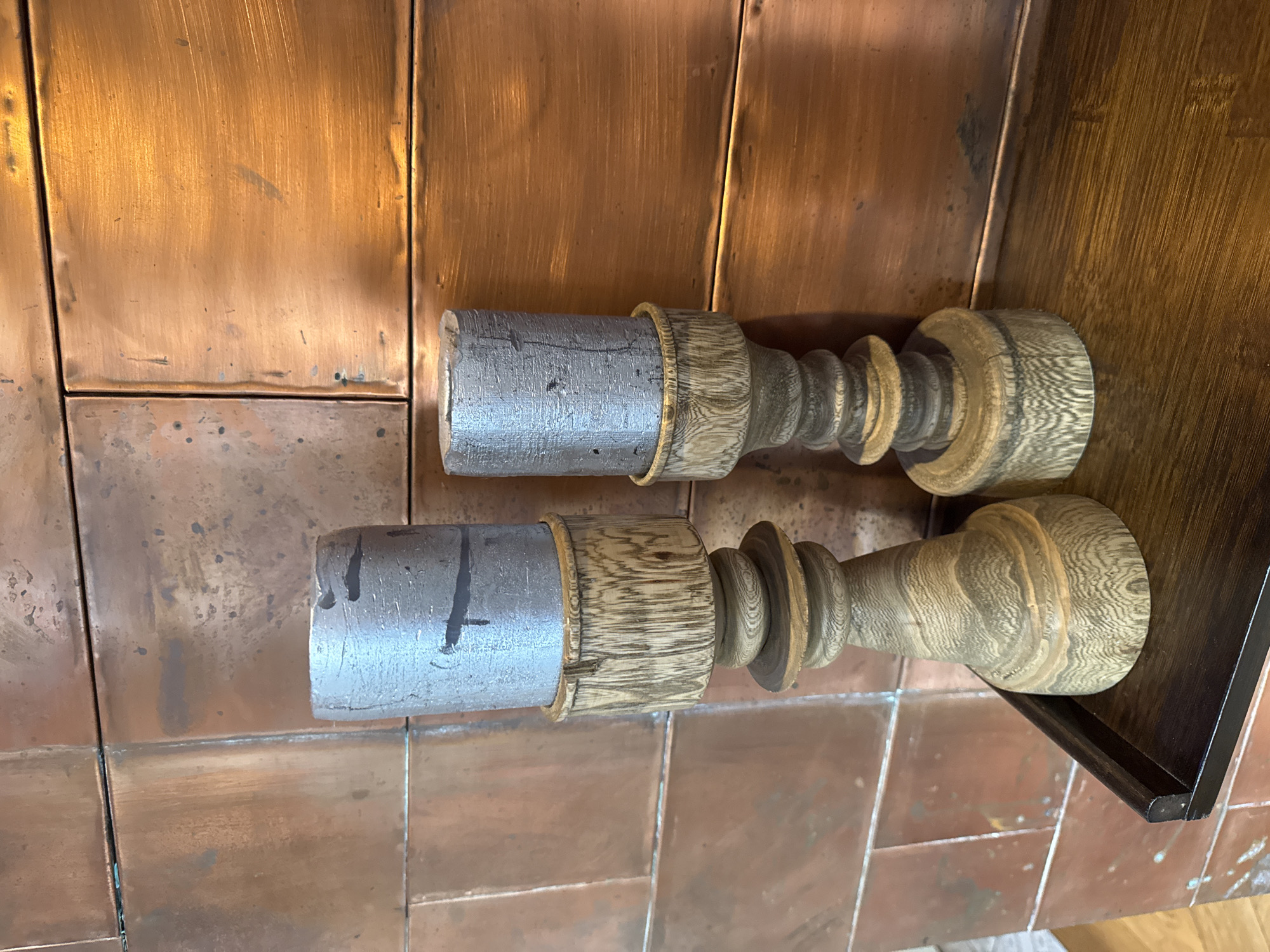Two wooden candle holders with metal bases are displayed on a tiled surface. The candle holders have a turned wood design with distinct rings and a natural wood finish that highlights the grain. The metal bases are cylindrical and appear to be made of a grey metal, possibly aluminum or steel, with visible tool marks and some black markings. The candle holders are positioned side-by-side and slightly angled. The background consists of copper-colored tiles and a dark wooden surface, suggesting an indoor setting. The lighting is warm and appears to be artificial, casting subtle shadows. There is no visible text in the image. The scene is static, with no apparent activity or event taking place, focusing solely on the objects. The setting, with its tiled surfaces and wooden elements, might be a kitchen, a workshop, or a decorative display area in a home. The overall impression is one of rustic or craftsman-style decor.