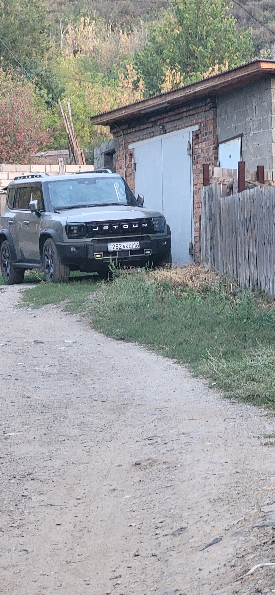 A gray, four-door SUV with a black grille and "JETOUR" branding is parked on a dirt road in front of a brick garage. The license plate reads "282 AKC 16". The garage has a large white door and a corrugated metal roof. A wooden fence is visible to the right of the garage. The background consists of a hill with sparse vegetation. The scene appears to be in a rural or semi-rural area of Oskemen, Kazakhstan, likely a residential property. No people are present.