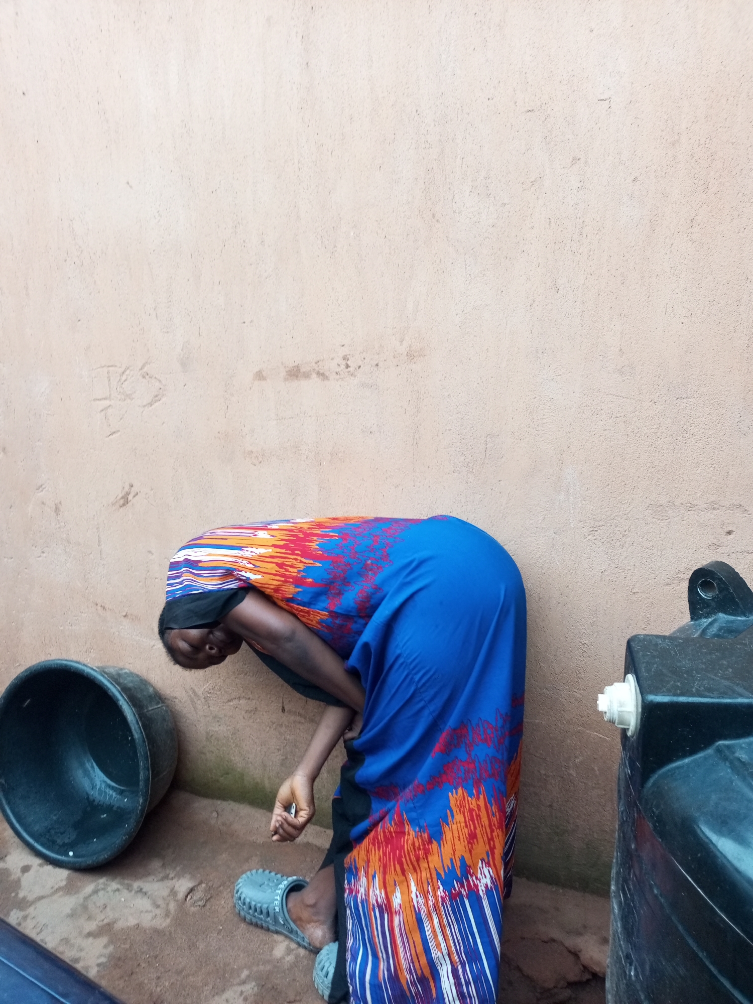 An unidentified person is bent over, with their head down, by a textured, tan-colored wall. The person is wearing a blue, orange, and white patterned dress. To the left of the person is a large, dark grey basin. To the right of the person, and partially obscuring the wall, is a black water tank with a white spigot. The ground appears to be dirt and sand, with some visible cracks. The scene is outdoors, with some moss or algae visible at the base of the wall. The lighting suggests it is daytime. The location is described as Tempure, Nigeria.
