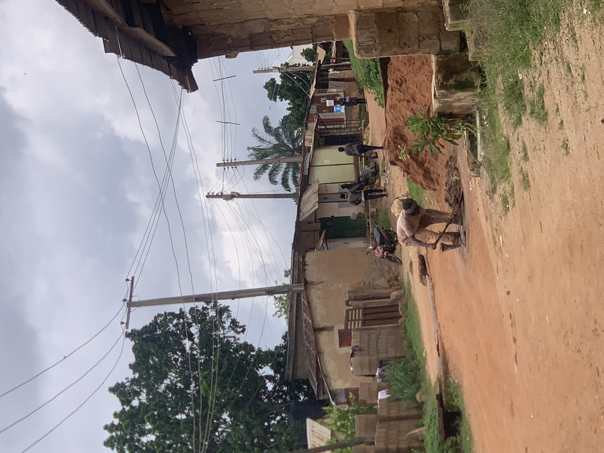 The image depicts a street scene in Jos, Nigeria, under a cloudy sky. The orientation of the image is sideways. In the foreground, a person is operating a tool, possibly for construction or repair work on the dirt road. Further down the street, several other individuals are visible, some walking and others gathered.
The setting includes a mix of simple buildings with corrugated metal roofs and some with brick walls. There are utility poles with overhead wires traversing the scene, suggesting the presence of electricity. Vegetation, including trees and grass, is scattered throughout, adding to the rural or semi-urban feel. The ground is predominantly reddish-brown dirt, indicative of the local soil.
The overall atmosphere appears to be one of daily life and ongoing activity. The time of day is indeterminate due to the cloudy weather, but the lighting suggests it is daytime. There are no discernible emotional expressions on the faces of the individuals, as they are either too far away or their features are not clear. No visible text is present in the image. The scene is characteristic of a developing area where infrastructure and daily routines are intertwined with the natural environment.