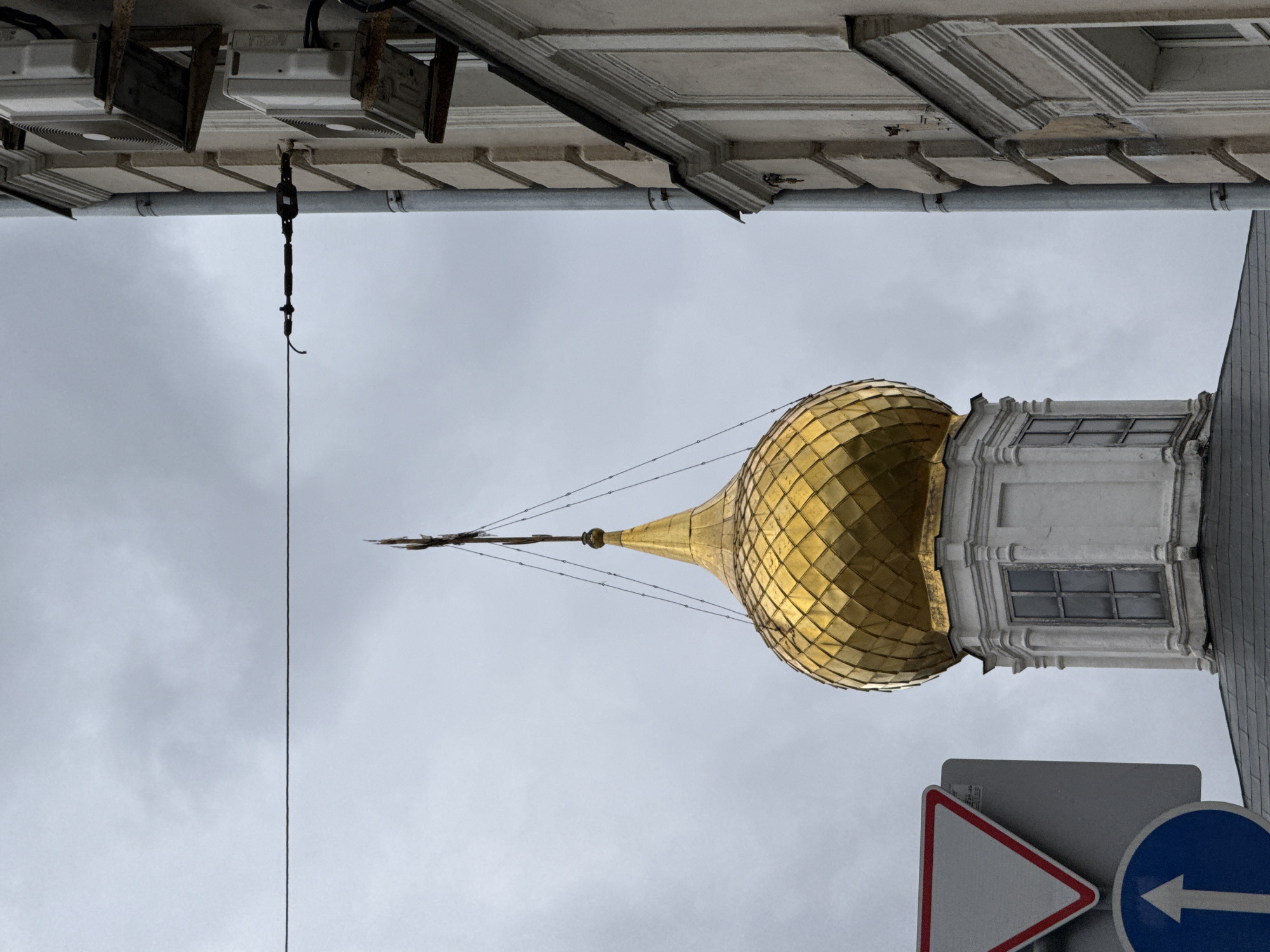 The image shows a close-up view of a golden dome of a church or cathedral, with a cross at its apex, set against an overcast sky. To the right, a portion of a light-colored building with architectural details and two outdoor air conditioning units is visible. In the foreground, on the left, a grey sign with a white triangle outlined in red, and a blue circular sign with a white upward arrow, are partially visible. The scene appears to be in an urban environment, possibly Moscow, Russia, due to the architectural style and the presence of traffic signs. The lighting is diffused, suggesting a cloudy or overcast day.