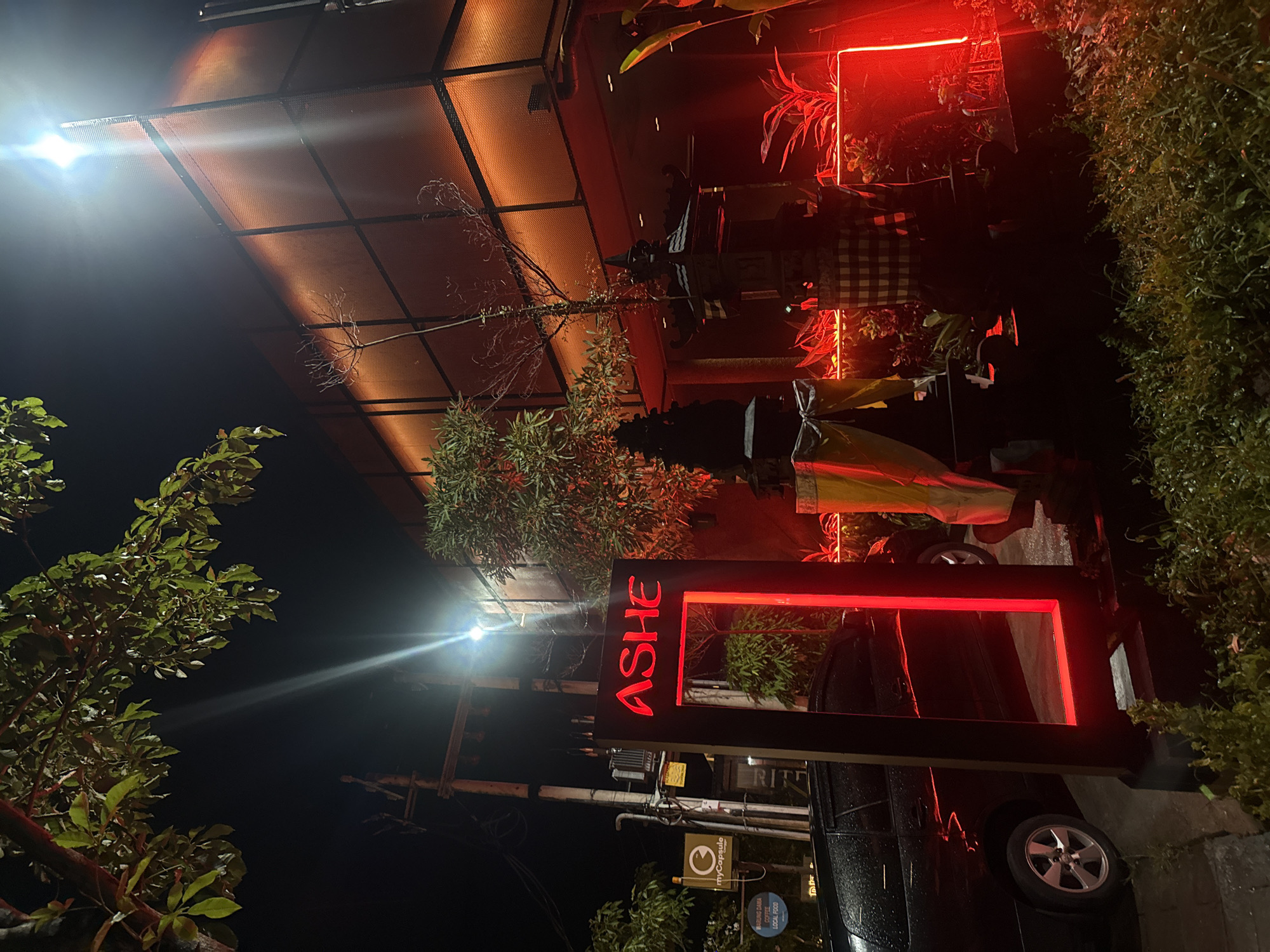 The image shows a nighttime scene in Unknown, Unknown. A red illuminated sign reads "ASHE" in bold letters. The sign is positioned near a dark-colored vehicle, possibly a car, with a visible tire. The environment features tropical greenery and a structure with a mesh-like roof. Decorations, including what appear to be traditional Balinese ornaments and flags, are present. A bright light source creates glare in the sky. Additional signs with logos are also visible.