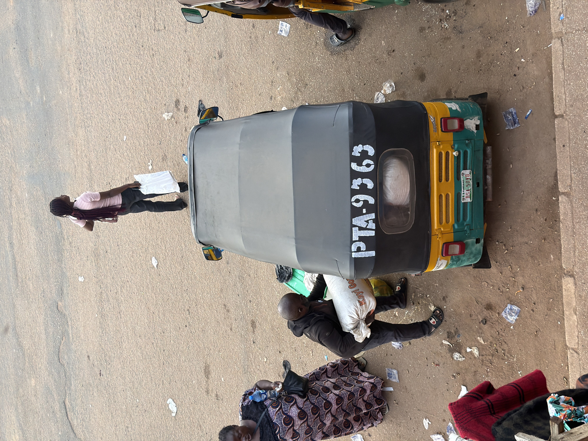 In Bukuru, Nigeria, an overhead view captures a street scene with several individuals and vehicles. A dark-colored auto-rickshaw, labeled with "PTA-9363" on its canopy and bearing the license plate "BSA-50YD," is parked prominently. A man is seen carrying a large sack towards the auto-rickshaw. To the left, a woman in a patterned dress stands with a water bottle and a handbag. Further to the left, another individual in dark clothing is visible. In the upper portion of the image, a person with long hair, wearing a pink shirt and dark pants, stands holding a white bag. Another auto-rickshaw, partially visible on the right, is also present, with a person seated inside. The ground is predominantly a light brown, sandy surface, littered with various small pieces of debris, including paper and plastic. The lighting suggests it is daytime, with a slightly overcast or hazy sky.