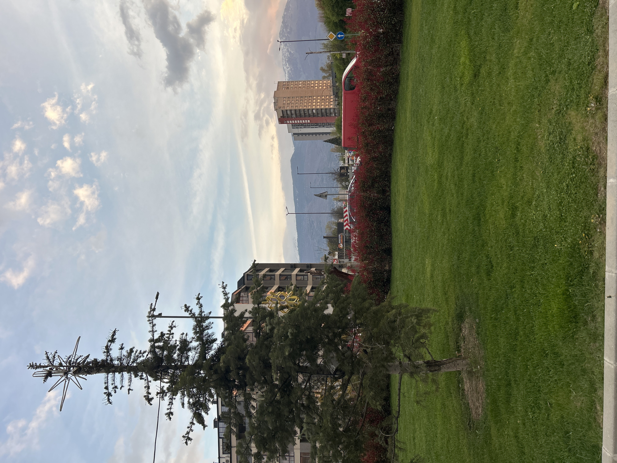 This image captures an urban landscape in Sofia, Bulgaria, showcasing a blend of natural elements and city infrastructure against a mountainous backdrop. The scene is depicted under a partly cloudy sky with soft, golden hues suggesting either late afternoon or early morning light.
In the foreground, a large, well-maintained expanse of green grass dominates the lower portion of the frame. To the left, a dense border of red-leafed bushes adds a splash of color, while a paved pathway runs along the right edge of the grassy area. A tall, slender coniferous tree, possibly a pine or fir, stands prominently on the right side of the green space. It is adorned with a large, elaborate star-shaped ornament at its top, hinting at a decorative or festive function.
Behind the foreground elements, several multi-story buildings are visible. One building stands out with numerous square windows, reflecting the ambient light. Other apartment-style buildings are partially obscured by the tree and bushes. A street appears to run between the green space and the buildings, featuring streetlights and various traffic signs, including a blue arrow and a yellow diamond sign. A section of a red vehicle, possibly a bus or truck, can be seen behind the red bushes.
The background is defined by the hazy, blue-grey outlines of distant mountains, stretching across the horizon. The sky above is a mix of light blue, white, and wispy grey clouds, with some areas touched by golden and pink tones.
No specific activity or event is taking place; the scene is static and serene. There is no visible text legible on any signs or buildings. The overall impression is one of urban tranquility, highlighting the integration of green spaces and city architecture with the natural mountainous environment of Sofia.