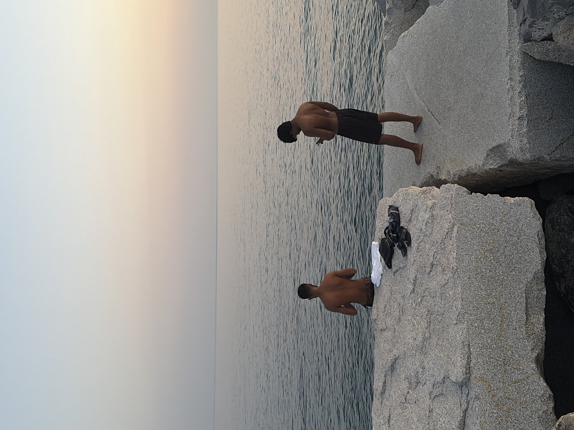 The image shows two young men standing on large granite rocks next to the sea. The scene is set during what appears to be late afternoon or early evening, as indicated by the soft, warm glow in the sky, transitioning from a pale yellow to a soft blue. The water is calm with gentle ripples. The man in the foreground is facing away from the camera, looking down at the water. He is wearing dark shorts. Behind him and slightly to the left, another man is also facing away from the camera, his head bent. He is wearing dark shorts as well. On the rock next to the man in the foreground, a white towel and a black bag are placed. The setting is a rocky coastline, possibly a breakwater or a natural formation of large boulders by the sea. The atmosphere appears serene and peaceful, with no text visible in the image. The location is identified as Imperia, Italy.