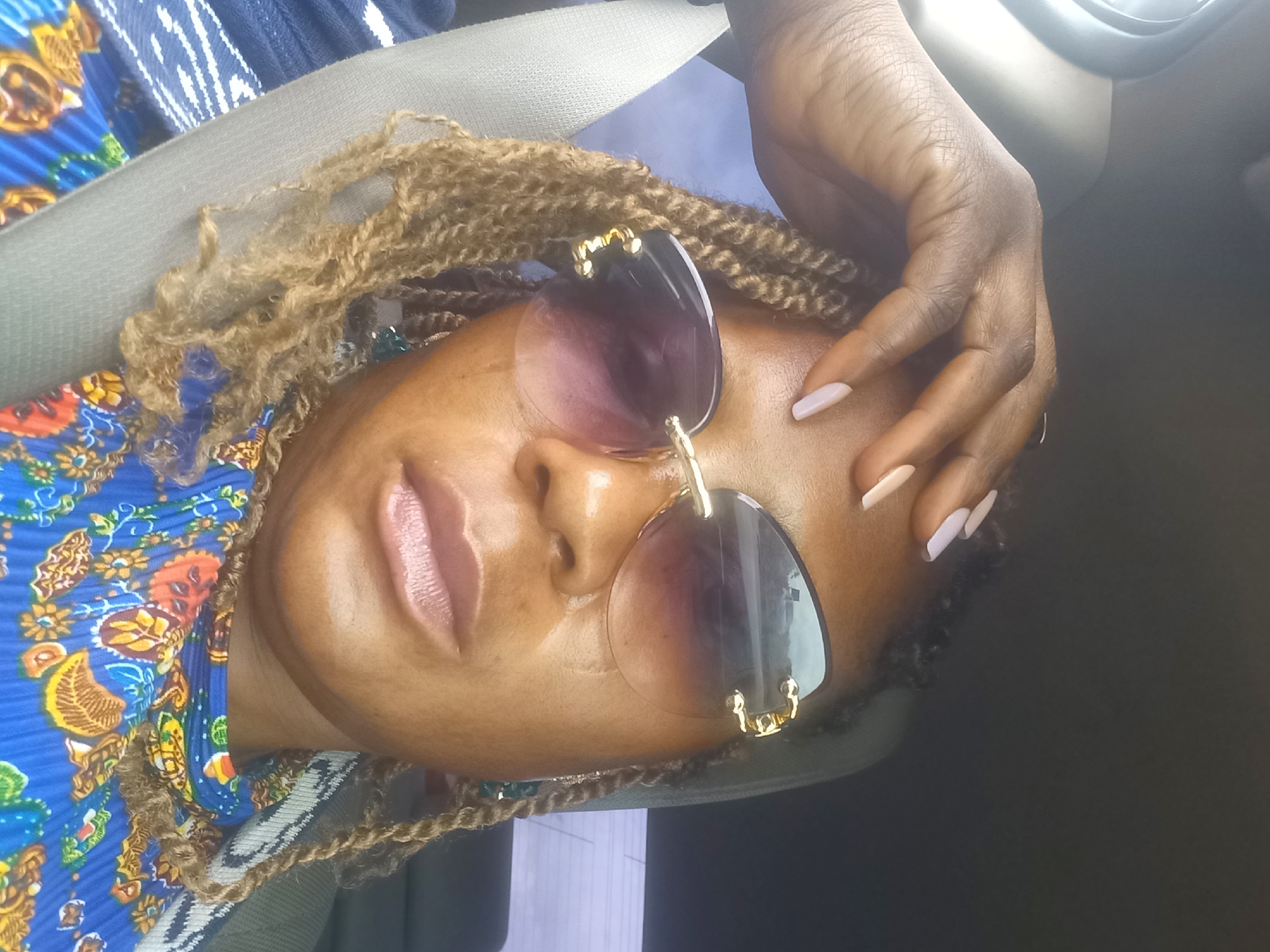 A woman with braided blonde hair and sunglasses is sitting in a car, with her hand on her forehead. She is wearing a blue and red patterned shirt, and a seatbelt is visible across her chest. The background shows the interior of a car and a blurred outdoor scene. The location is specified as Mangu Halle, Nigeria.