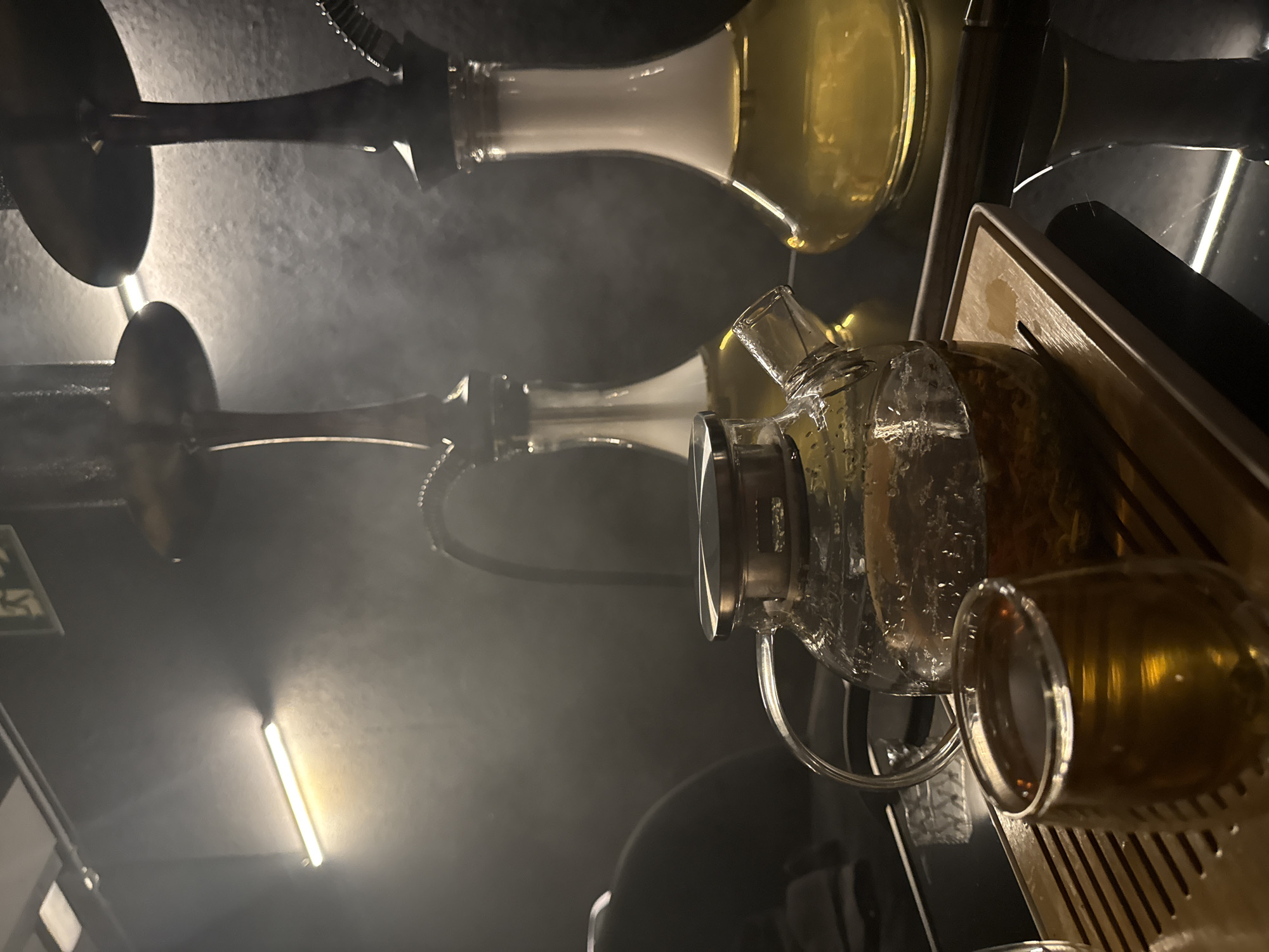 The image depicts an indoor setting, likely a lounge or bar, with a dark ambiance illuminated by warm lighting. Two ornate hookahs with golden accents are visible in the background, with wisps of smoke creating a hazy atmosphere. In the foreground, a clear glass teapot filled with a golden liquid and tea leaves sits on a wooden tray. Next to it, a small glass cup also contains the amber-colored beverage. The overall scene suggests a relaxed and possibly social gathering focused on enjoying tea and hookah. No specific landmarks or elements suggest the location is Barcelona, Spain.