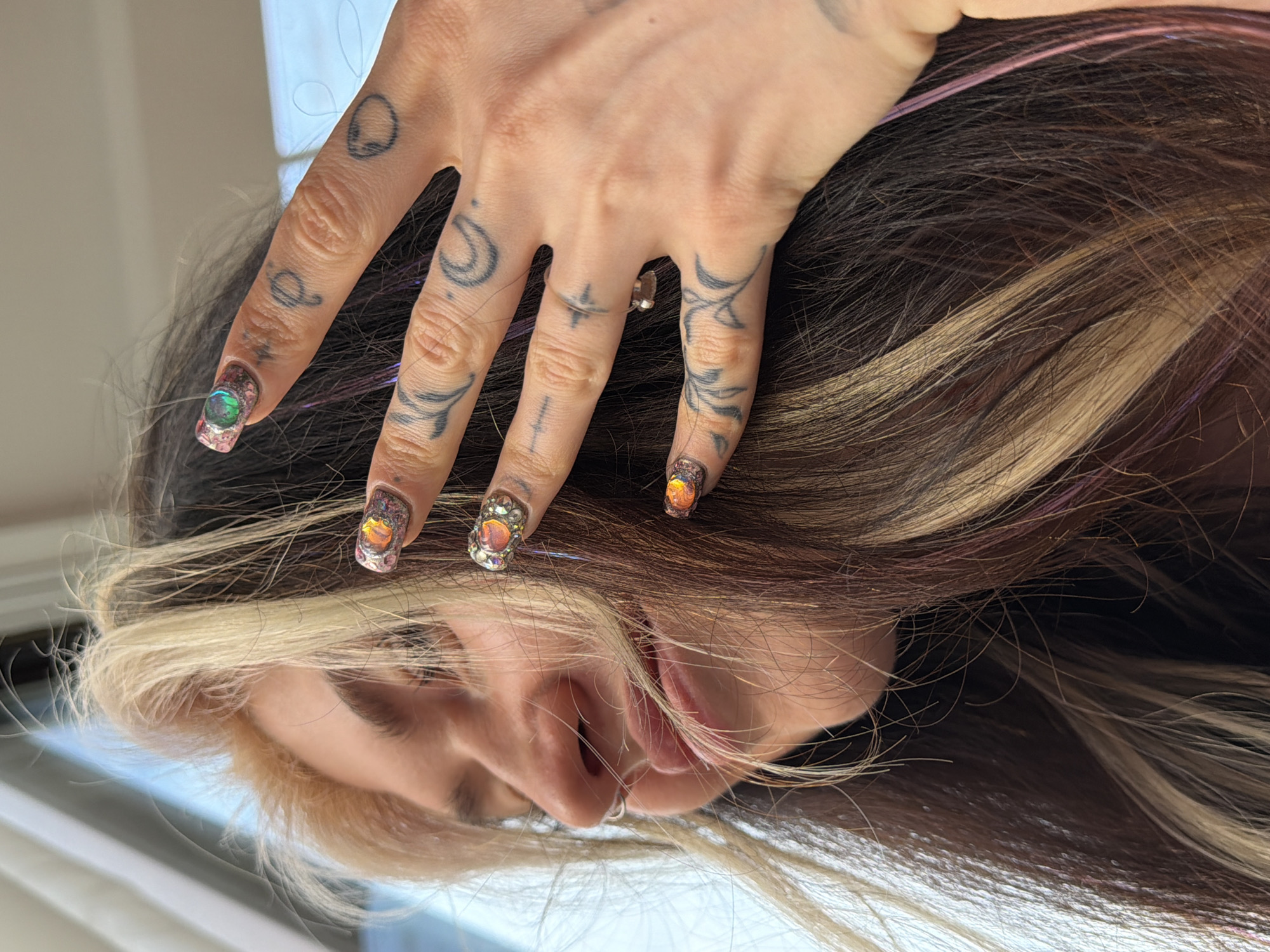 A woman with brown and blonde hair lies sideways. Her hand, adorned with tattoos and elaborate nail art, rests on her hair. The nail art features decorative jewels. A silver ring is visible on her finger. She has a nose ring. The setting appears to be indoors. While the image provides personal details about the subject, it does not provide any information regarding a specific location in Budva, Montenegro.