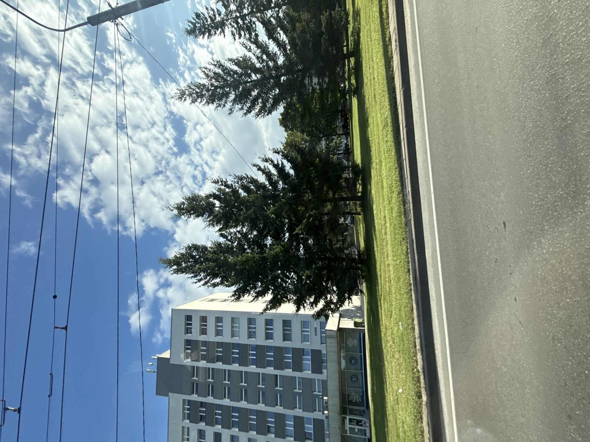 In Tallinn, Estonia, a street view captures a multi-story apartment building with a grey and white facade and numerous windows. In front of the building, a well-maintained lawn is interspersed with several tall pine trees. Overhead, power lines crisscross the sky, supported by a lamppost visible on the right. The foreground shows a paved road with a white line marking the lane. The sky is blue with scattered white clouds.