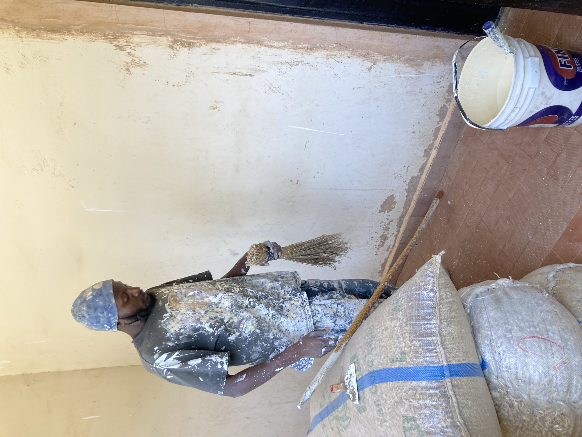 A person is painting a wall. The person is wearing paint-splattered clothing and a hat. They are holding a brush in their left hand. To their right are bags and a paint bucket. The location is Jos, Nigeria.