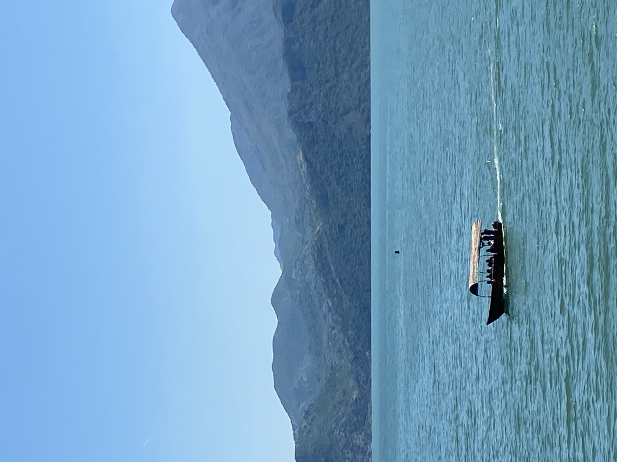 The image depicts a long, narrow boat carrying several people on a body of water, presumably a lake, in Kruševica, Montenegro. Mountainous terrain is visible along the shoreline, with a clear blue sky above. A small, dark object, possibly a buoy or marker, floats near the boat in the water. The overall scene is serene and outdoorsy.
