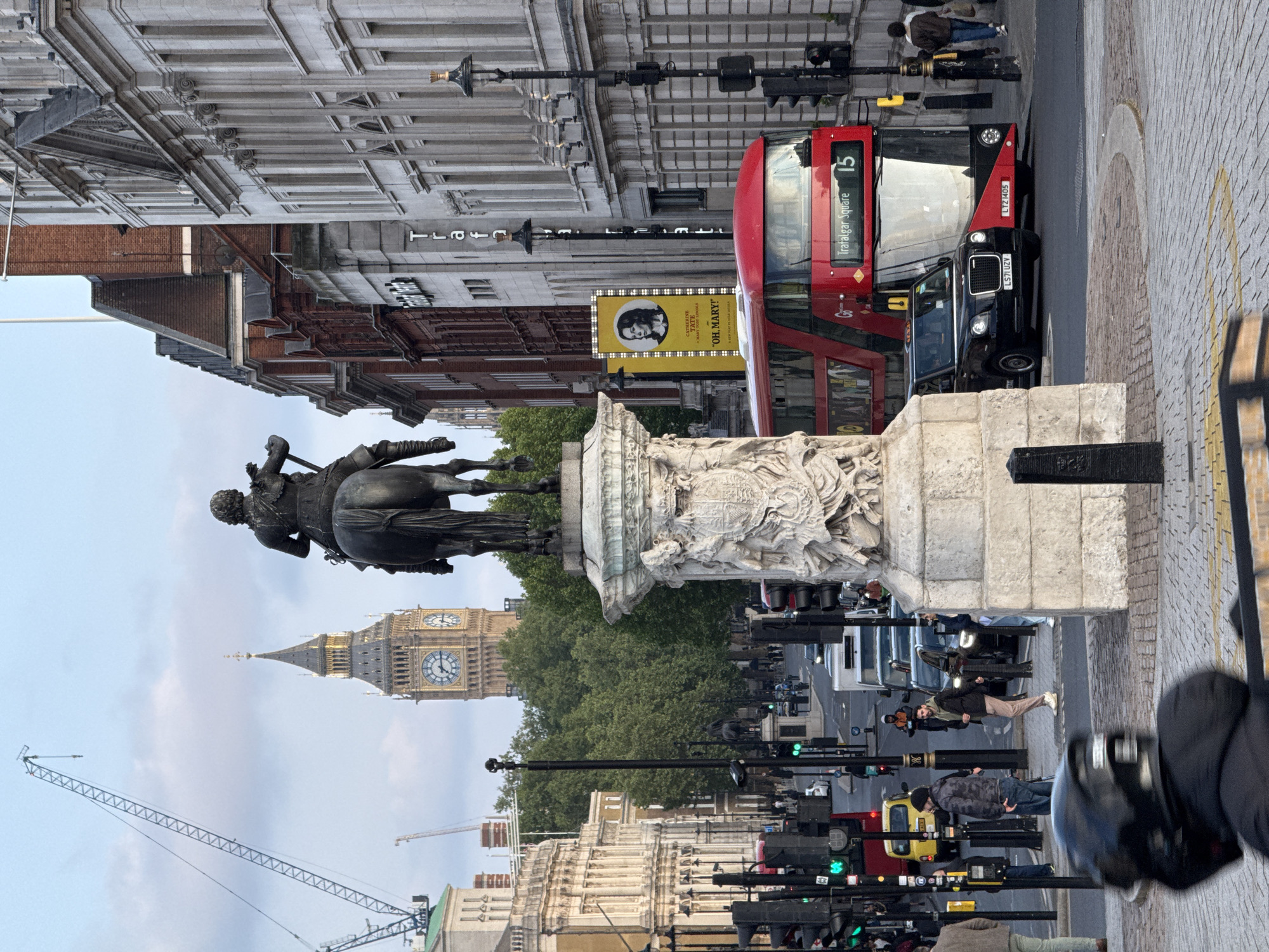 This image showcases a street scene in London, United Kingdom. At the forefront is a statue of a man on horseback, atop an ornate stone pedestal. Behind the statue is the iconic Big Ben clock tower, partially obscured by trees. A red double-decker bus, identified by "Trafalgar Square 15", and a black taxicab are visible on the street, with registration plates LTZ 1405 and LS7I UZV. A yellow advertisement banner shows a person's face and the text "OH, MARY!". The street is bustling with pedestrian traffic, and traffic lights line the road. A large crane is visible in the background. The buildings are a mix of old and modern architecture.